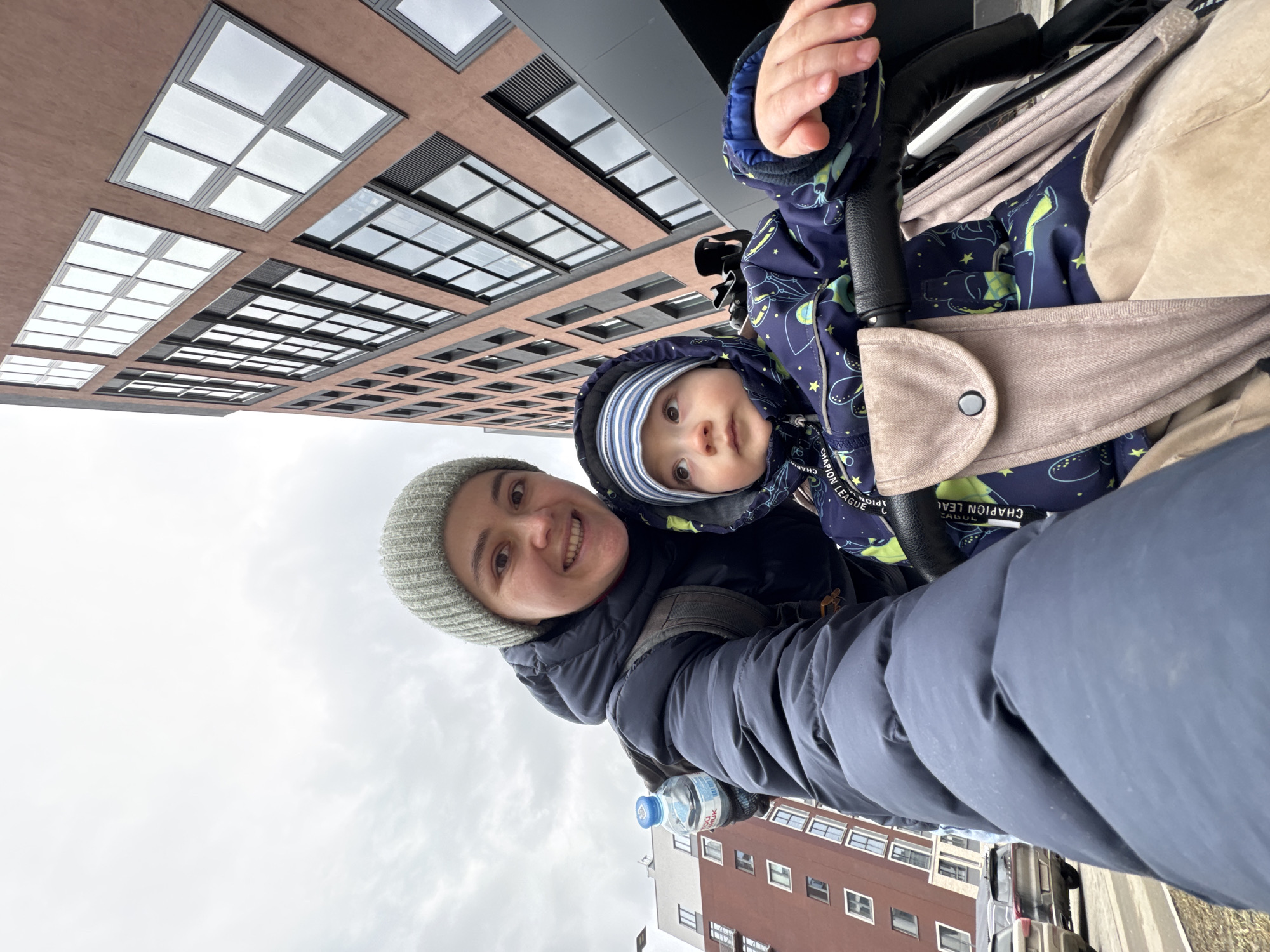 A woman in a gray knit hat and navy jacket smiles for a selfie with a baby in a blue astronaut-print snowsuit in a stroller. The photo is taken from a low angle looking up at a modern building with grid-like windows. A water bottle is visible in the woman's hand. The building is located in Izhevsk, Russia.