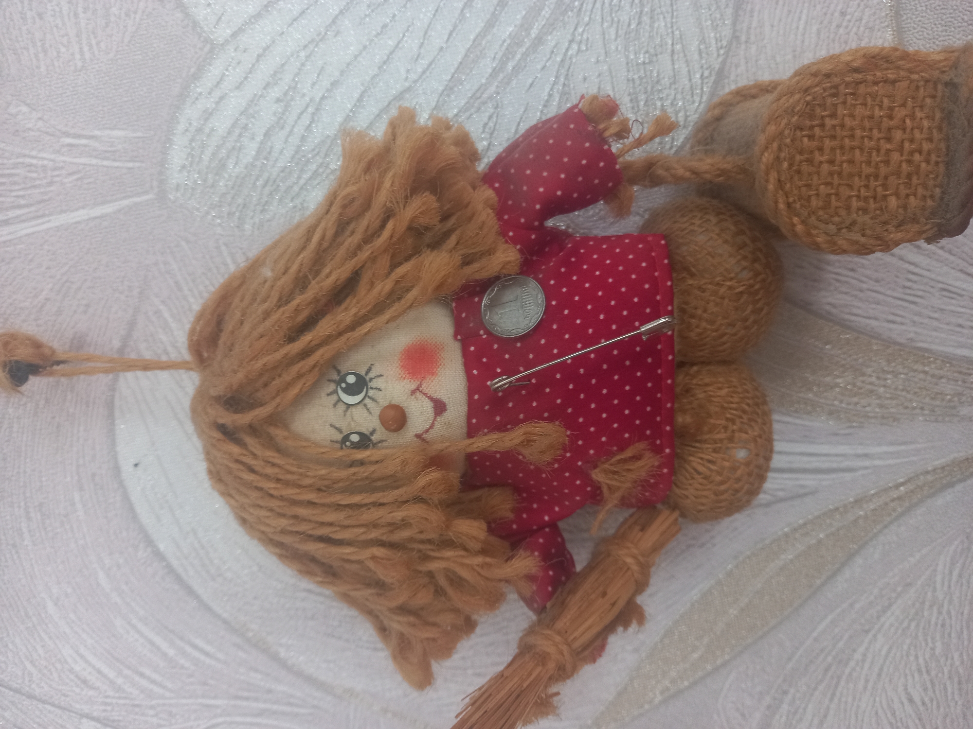 A handmade doll, possibly a witch, hangs against a textured white wallpaper. The doll has a red polka-dot shirt and a broomstick. It has yarn hair and a coin pinned to its shirt. The doll is suspended by a string from above. The wallpaper has a floral or leaf-like pattern.