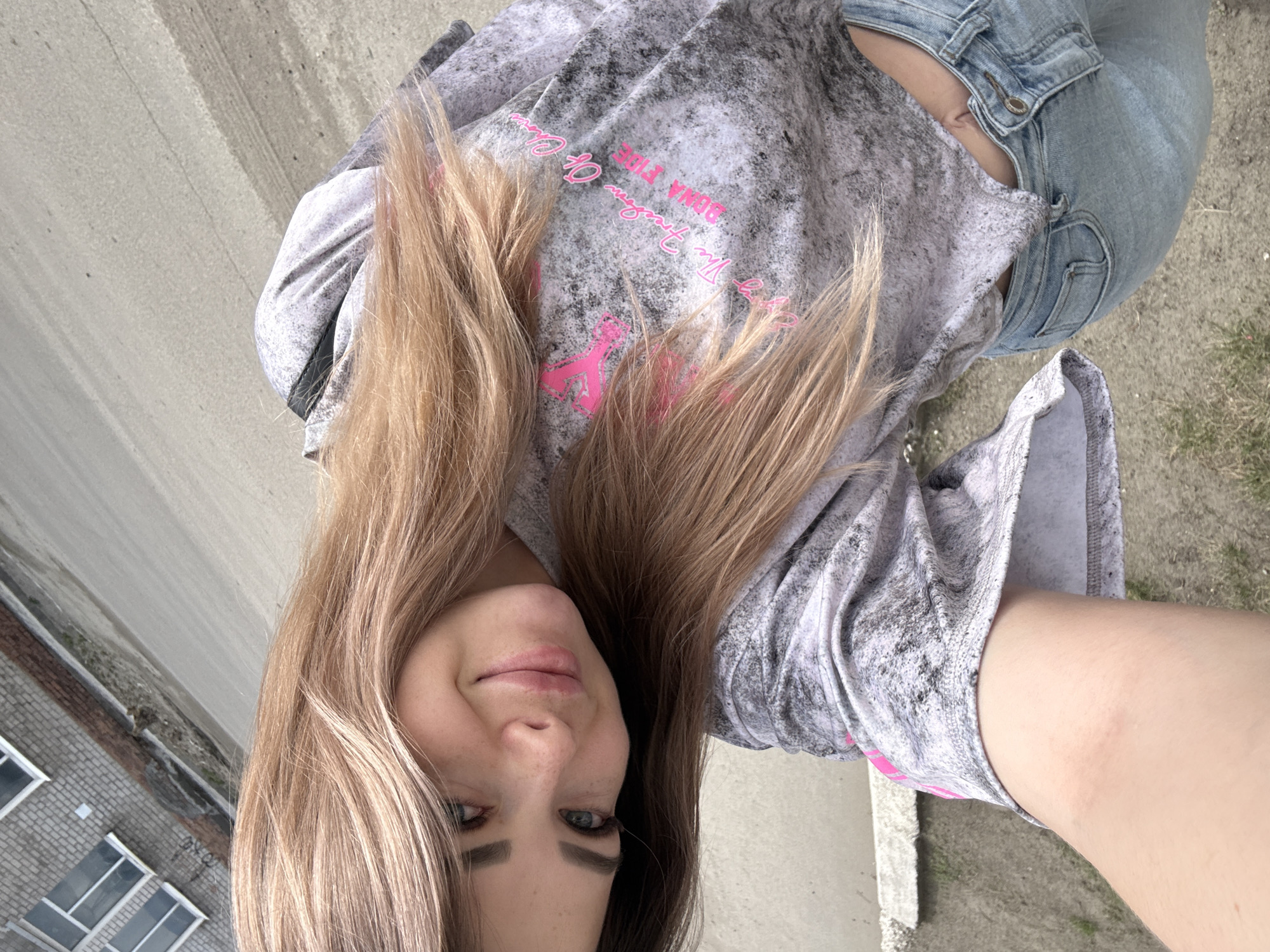 A young woman is taking a selfie. She has long blonde hair and is wearing a gray and white tie-dye sweatshirt with pink writing. She is also wearing blue jeans. The background is blurry, but it appears to be a city street with a brick building on the left.