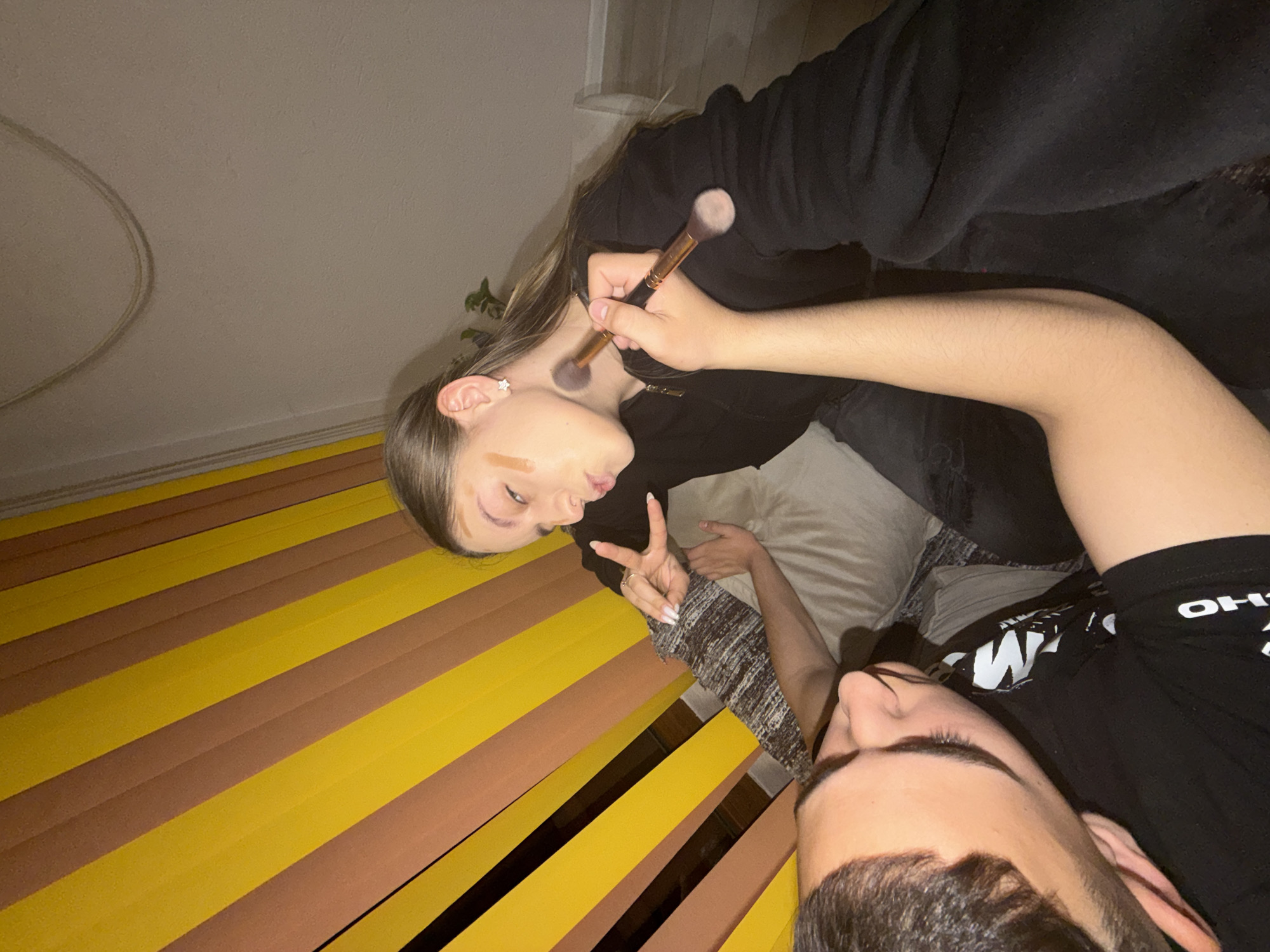 Two individuals are present in the image. One person is applying makeup to the face of the other with a makeup brush. The individual receiving makeup is reclining with their hand raised in a peace sign. The background features a yellow and brown striped surface, and a neutral-colored wall. The location cannot be determined to be Panagyurishte, Bulgaria from the given image.