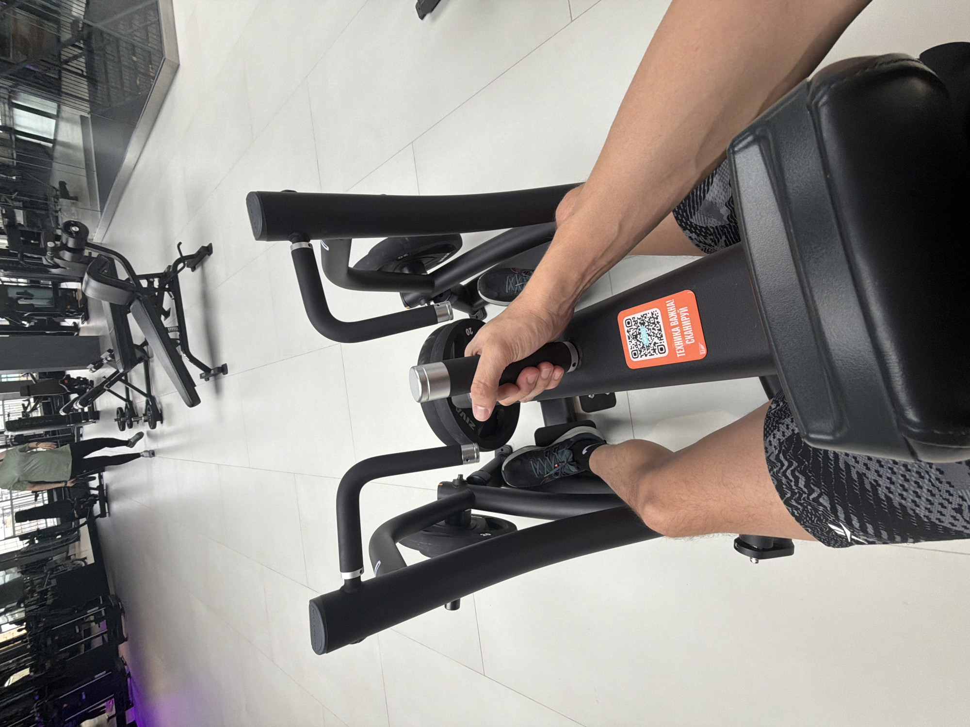 A person is using a weight machine in what appears to be a gym setting. The person is grasping the handle of the machine with their right hand. The lower part of their body is visible, wearing patterned shorts and athletic shoes. The weight machine has various black components, including handles, bars, and weight plates. There is an orange sticker with a QR code on the machine. In the background, there are more gym equipment and people exercising. The scene is likely indoors with a tile floor. The location is a gym in Krasnoyarsk, Russia.