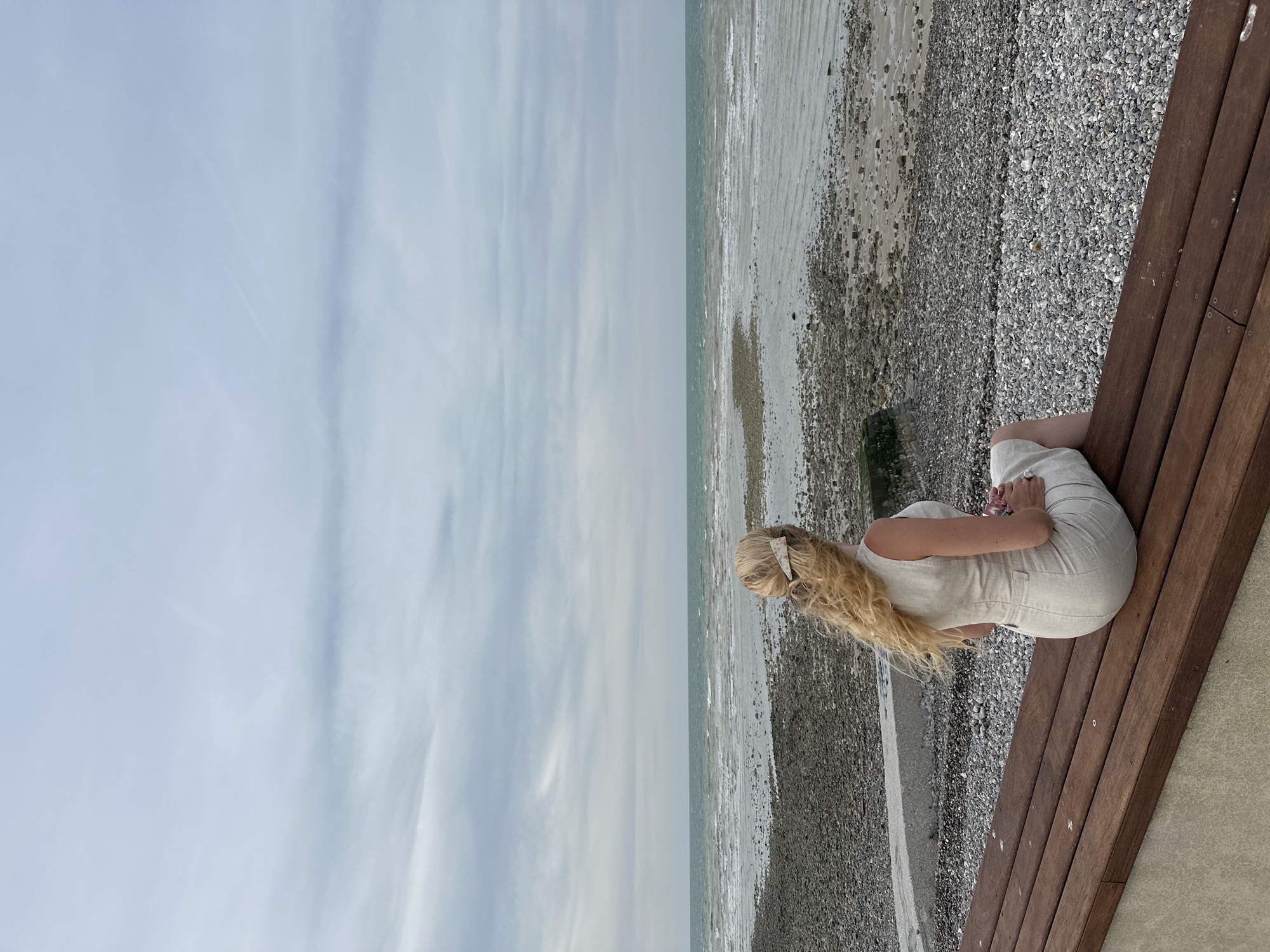 A young woman with long blonde hair, wearing a light-colored sleeveless dress, is sitting on a wooden bench facing the sea. She has a hair clip in her hair and is looking out at the water. The beach is stony, and the sea is calm with small waves. The sky is overcast. The location is the beach in Saint-Valery-en-Caux, France.