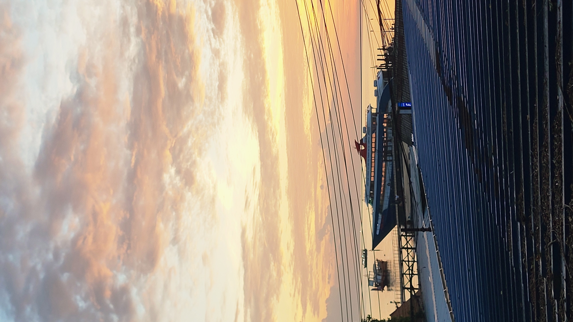 The image depicts a sunset scene over a body of water, likely in the Ko Pha-ngan District of Thailand. In the foreground, a dark, corrugated metal roof structure dominates the lower portion of the frame. Above this roof, a series of dark power lines stretch horizontally across the image, partially obscuring the view of the water and sky.
On the water, a sleek, modern ferry with a red and white superstructure is docked. It appears to be a passenger vessel, with numerous windows visible along its sides. To the left of the ferry, a smaller, older wooden boat with a covered cabin is also present. Both boats are moored near a wooden pier structure.
The sky above is filled with dramatic, colorful clouds, illuminated by the setting sun. Hues of orange, pink, and yellow are mixed with softer gray and white tones, indicating a vibrant twilight. The horizon line between the water and the sky is visible, with the sun casting a warm glow. The overall impression is one of a coastal or harbor area during the evening. No people are clearly visible in the scene.