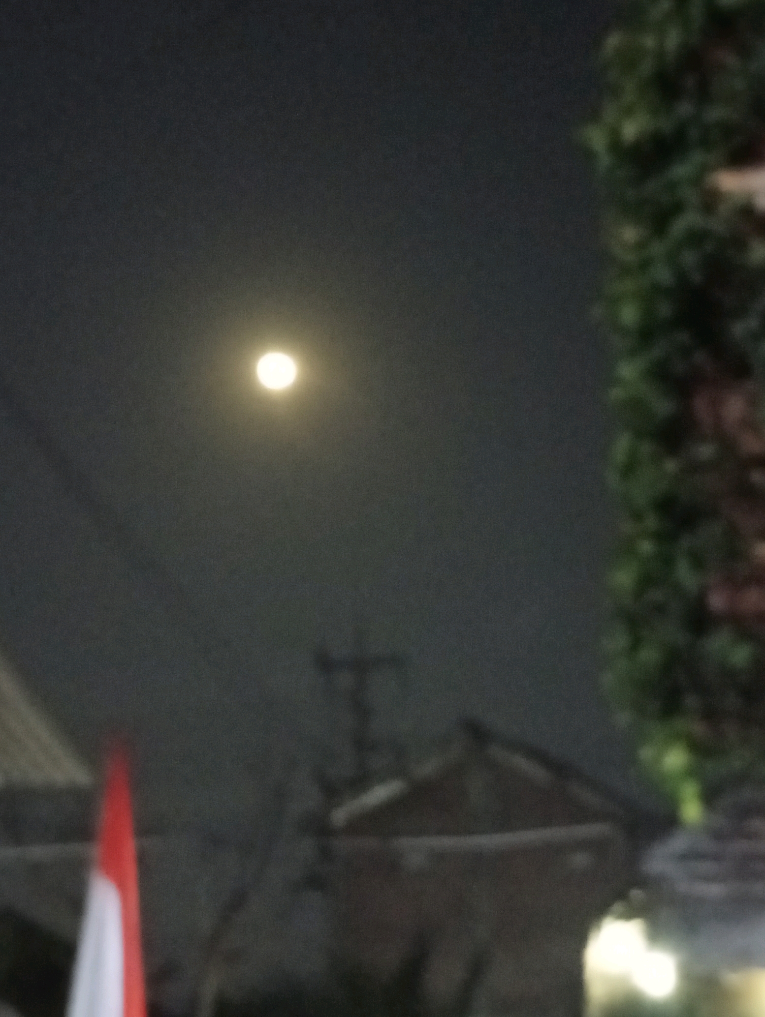 The image shows a night scene with a full moon in a dark sky. In the foreground, there are hints of buildings, a flag that appears to be red and white, and a tall, leafy plant on the right side. The overall impression is slightly blurry, suggesting a low-light or handheld photograph. The location is specified as Kediri City, Indonesia.
