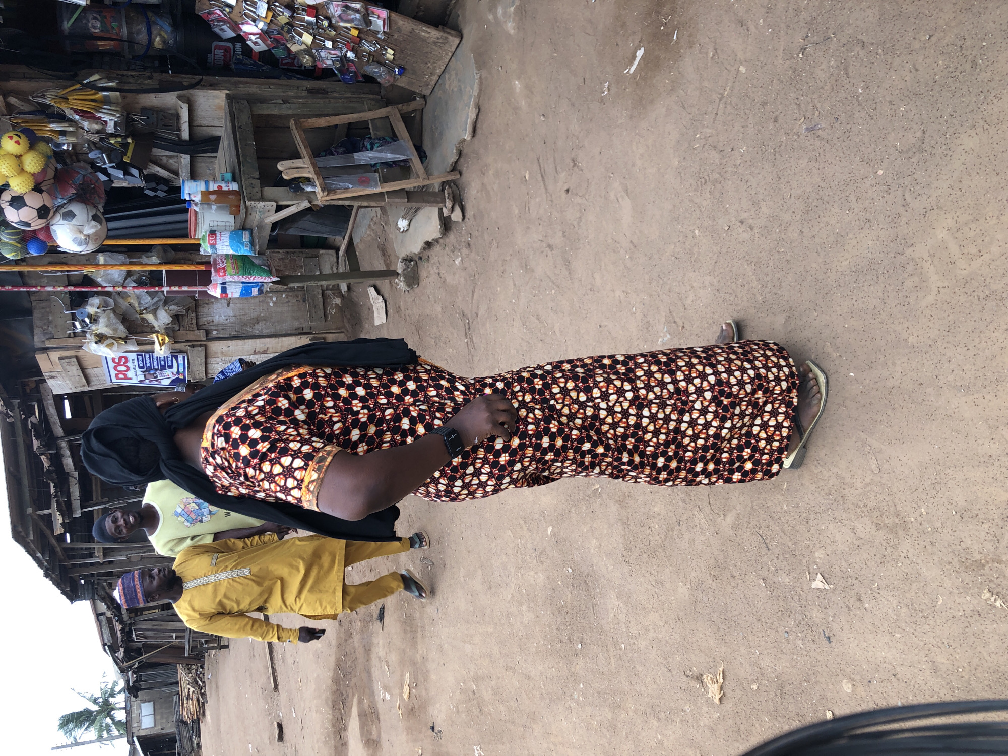 In Ijoko, Nigeria, a woman wearing a black headscarf and a patterned dress stands in a dusty outdoor market. She is facing away from the camera, her right hand resting on her hip, where a black smartwatch is visible. To her left, two men are walking. One man, in the foreground, wears a yellow traditional outfit and a cap. Behind him, another man in a light yellow t-shirt and a dark cap smiles at the camera. The background reveals the market stalls with wooden structures and various goods displayed, including cleaning supplies, locks, and other items. The ground is unpaved and dusty, typical of an outdoor market in the region.