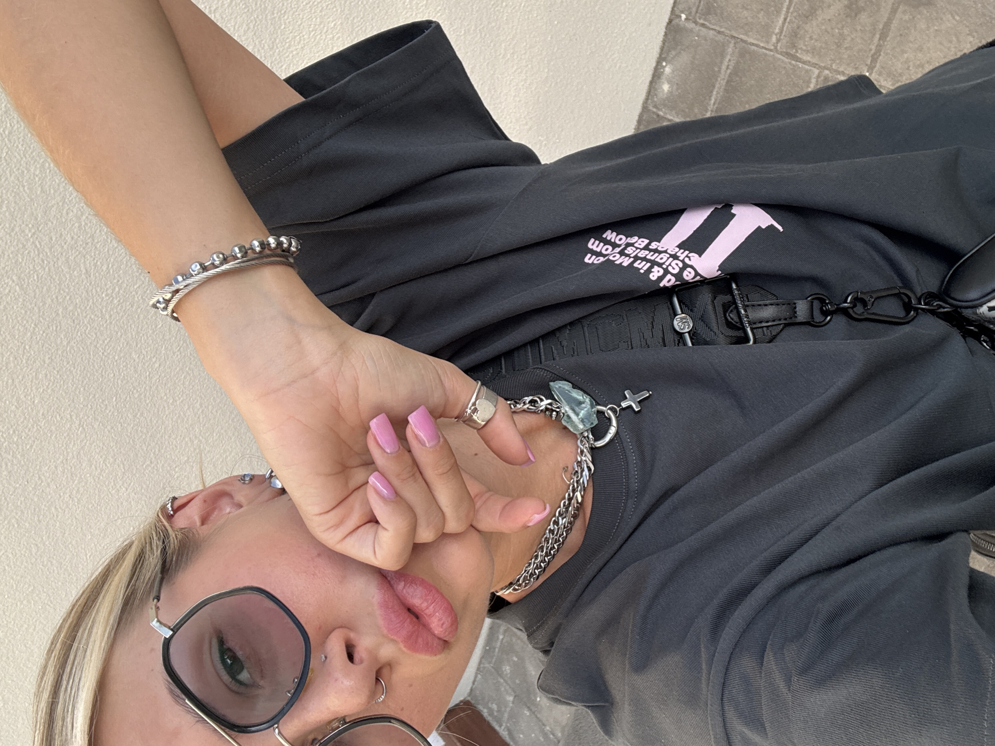 A young woman with blonde hair, wearing oversized sunglasses and a dark grey t-shirt, poses for a selfie. She has several piercings in her ears and nose, and her fingernails are painted a light pink. She is wearing a silver bracelet and necklace with a cross pendant. A black strap with a carabiner is visible on her t-shirt. The background is a plain, textured wall. The lighting suggests it is daytime.