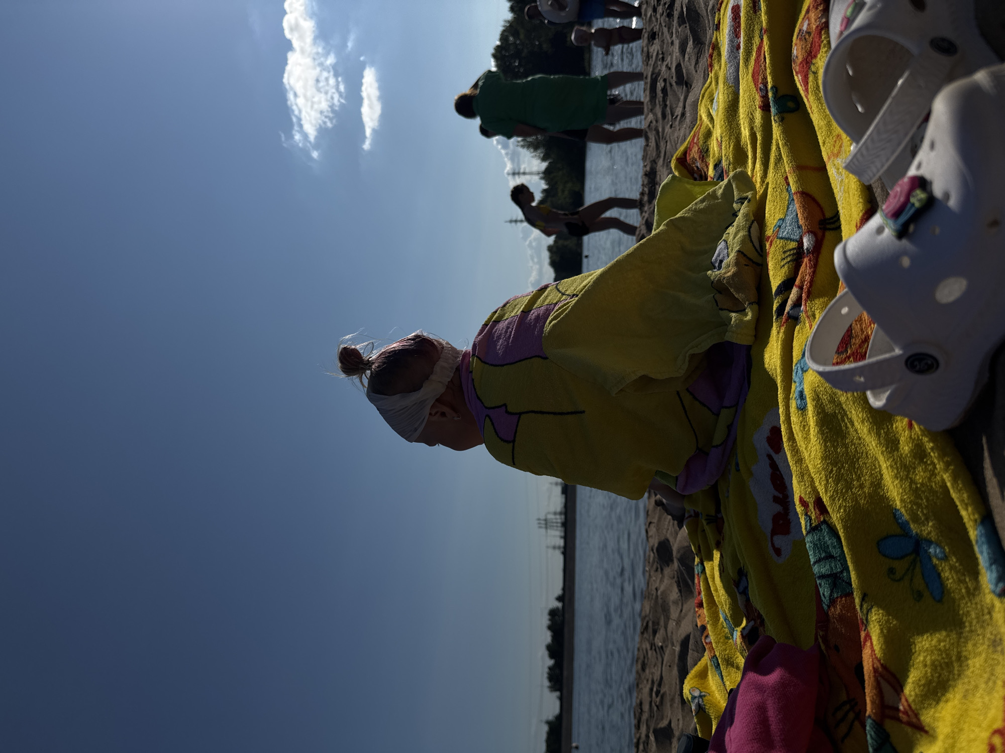A child is seated on a colorful beach towel on a sandy beach, facing away from the camera and towards a body of water. The child is wrapped in a yellow towel with purple and pink accents, and a white band is around their head. The beach towel features a pattern of various cartoon characters. In the foreground, a pair of white Crocs are visible. In the background, several people are standing near the water, some of whom appear to be swimming. The water is calm, and the sky is clear blue with a few scattered clouds. The scene is illuminated by bright sunlight. This scene is likely taking place at a beach located in or near the city of Noviy, Russia, given the context provided.