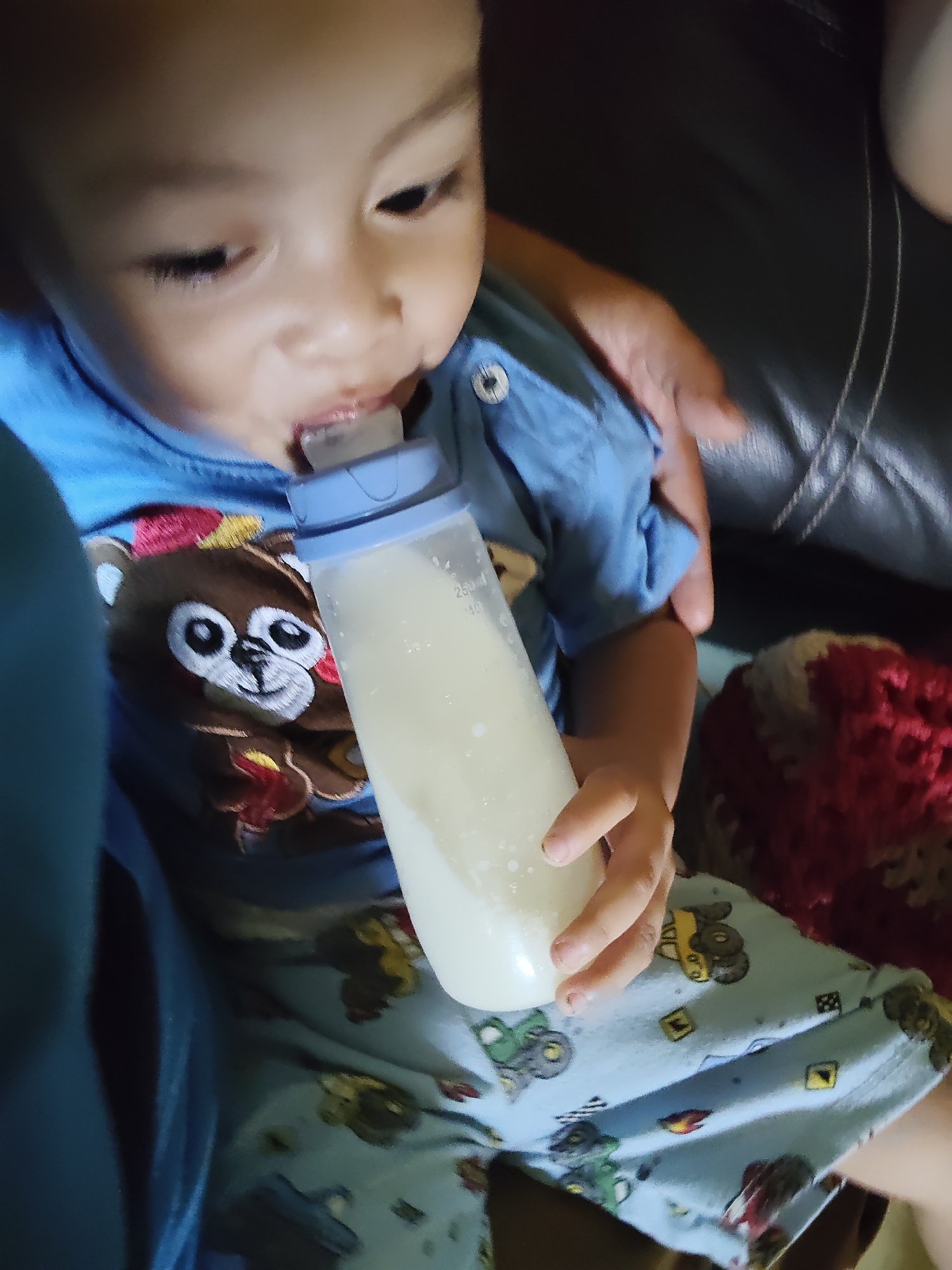 A young child, likely a toddler, is holding and drinking from a baby bottle. The child is wearing a blue t-shirt with a cartoon bear design and blue pajamas with vehicle prints. The background appears to be a dark, possibly leather, surface, such as a couch, with some red and beige fabric, possibly a blanket, visible on the right side. The scene suggests a domestic setting. There is no information in the image that would allow for determining its location within Tanah Grogot, Indonesia.