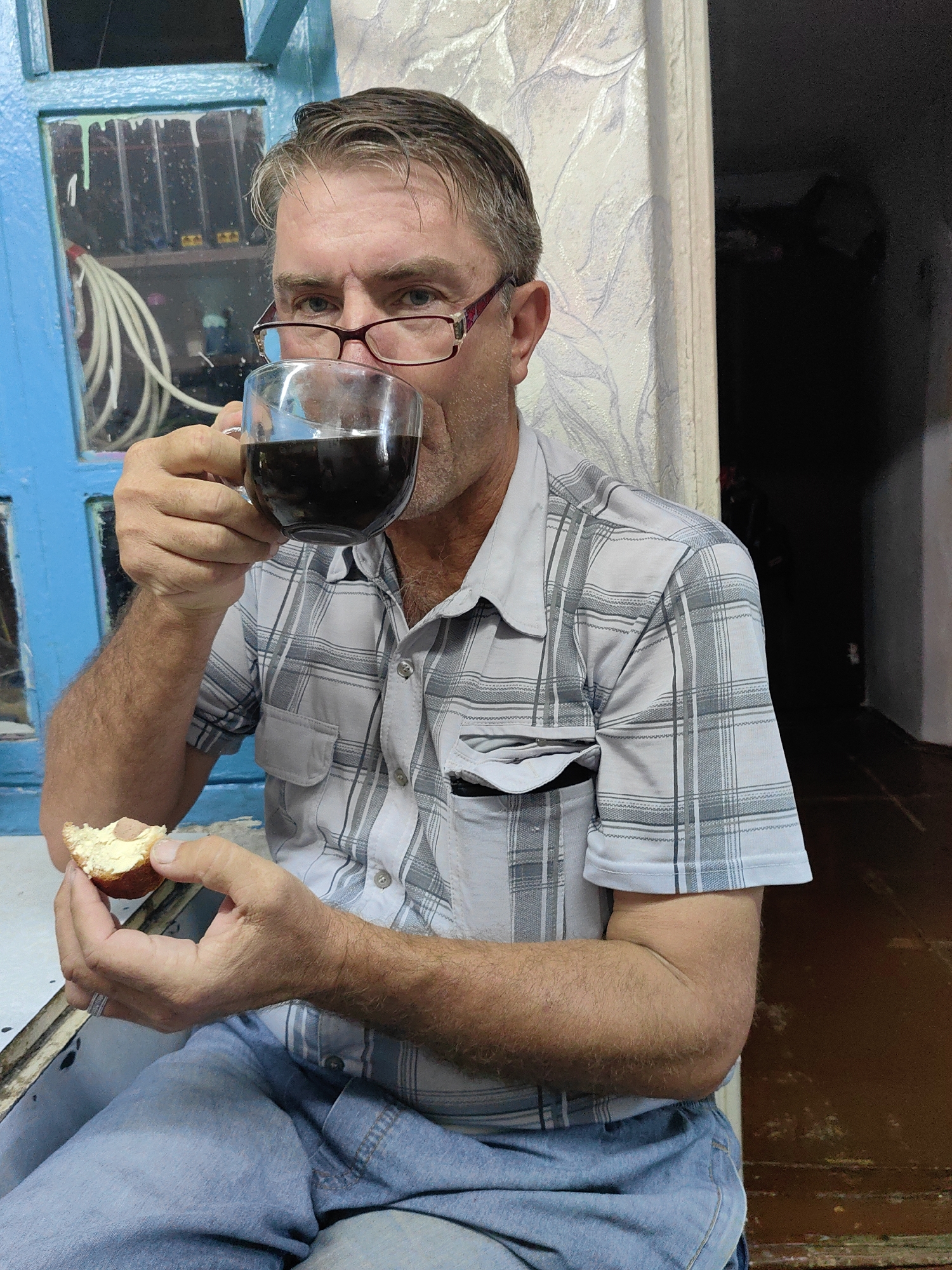A middle-aged Caucasian male with short, graying hair and a light beard is holding a glass mug of dark liquid to his lips with his right hand and a piece of bread with a pink sausage slice on top in his left hand. He is wearing a light-colored, checkered short-sleeved shirt and blue jeans. He has on reddish-rimmed glasses. The man is seated, and his left arm is resting on a surface. In the background, there is a blue-painted window with glass panes and a light-colored, patterned wallpaper. To the right of the man, a dark, unlit doorway or hallway is visible. The overall setting appears to be an indoor space, possibly a room or a porch, in the city of Shymkent, Kazakhstan.