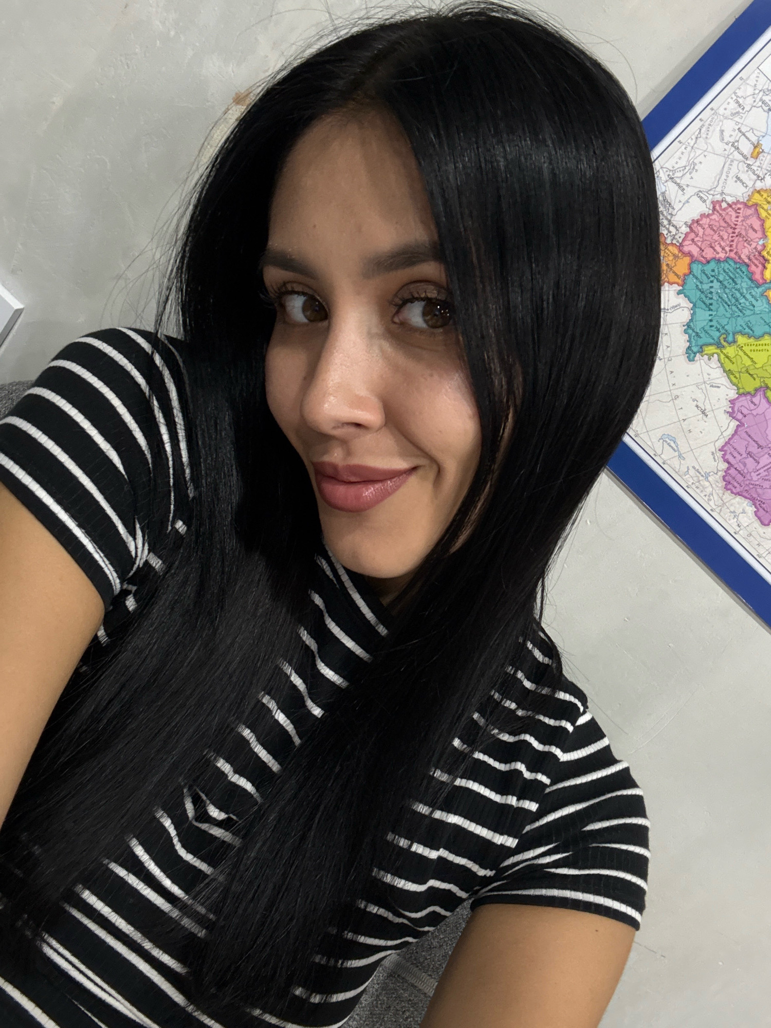 The image is a close-up selfie of a woman with long, dark hair. She is wearing a black and white striped shirt. Her head is tilted to her left, and she is looking towards the camera with a slight smile. Her makeup includes mascara and lipstick. In the background, a map is visible on the wall, featuring colorful regions and text in a Cyrillic alphabet. To the left of the woman, a portion of a light-colored wall is visible. No specific location in Votkinsk, Russia is identifiable from the image.