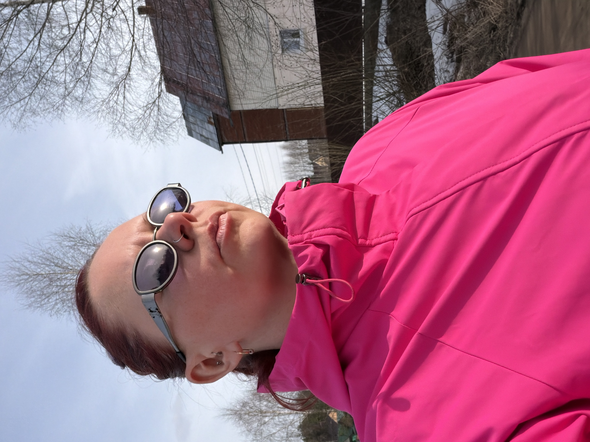 A woman wearing sunglasses and a bright pink jacket stands outdoors in Chaikovsky, Russia. Behind her is a two-story building with a reddish-brown, sloped roof and white siding on the upper level, with darker siding on the lower level. Bare trees are visible in the background against a pale blue sky.