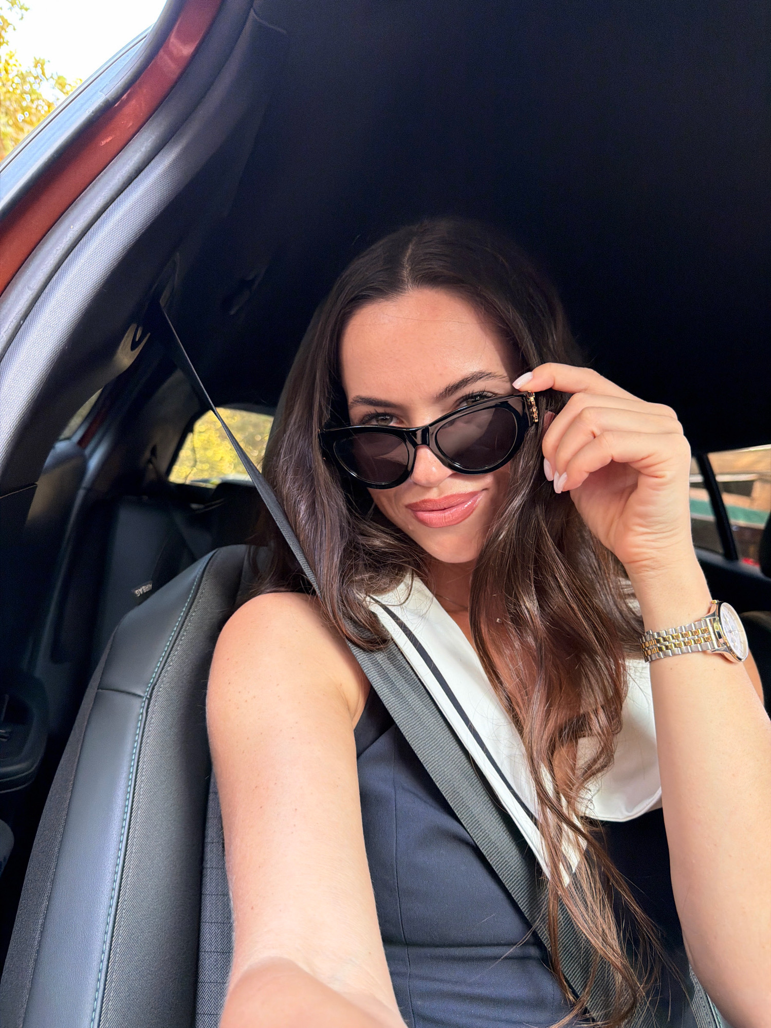 A woman with long, dark brown hair and light skin is sitting in the driver's seat of a car. She is wearing black, cat-eye sunglasses, a navy blue dress with a white collar, and a gold and silver watch on her left wrist. She is holding the arm of her sunglasses with her right hand. The interior of the car appears to be dark gray leather. A seatbelt is visible across her chest. Outside the car window, trees with yellow leaves can be seen, suggesting it is autumn. The image is taken from a slightly high angle, looking down at the woman. The overall scene is well-lit, likely by natural daylight. There are no identifiable landmarks or contextual clues in the image that would place the location specifically in Salou, Spain.