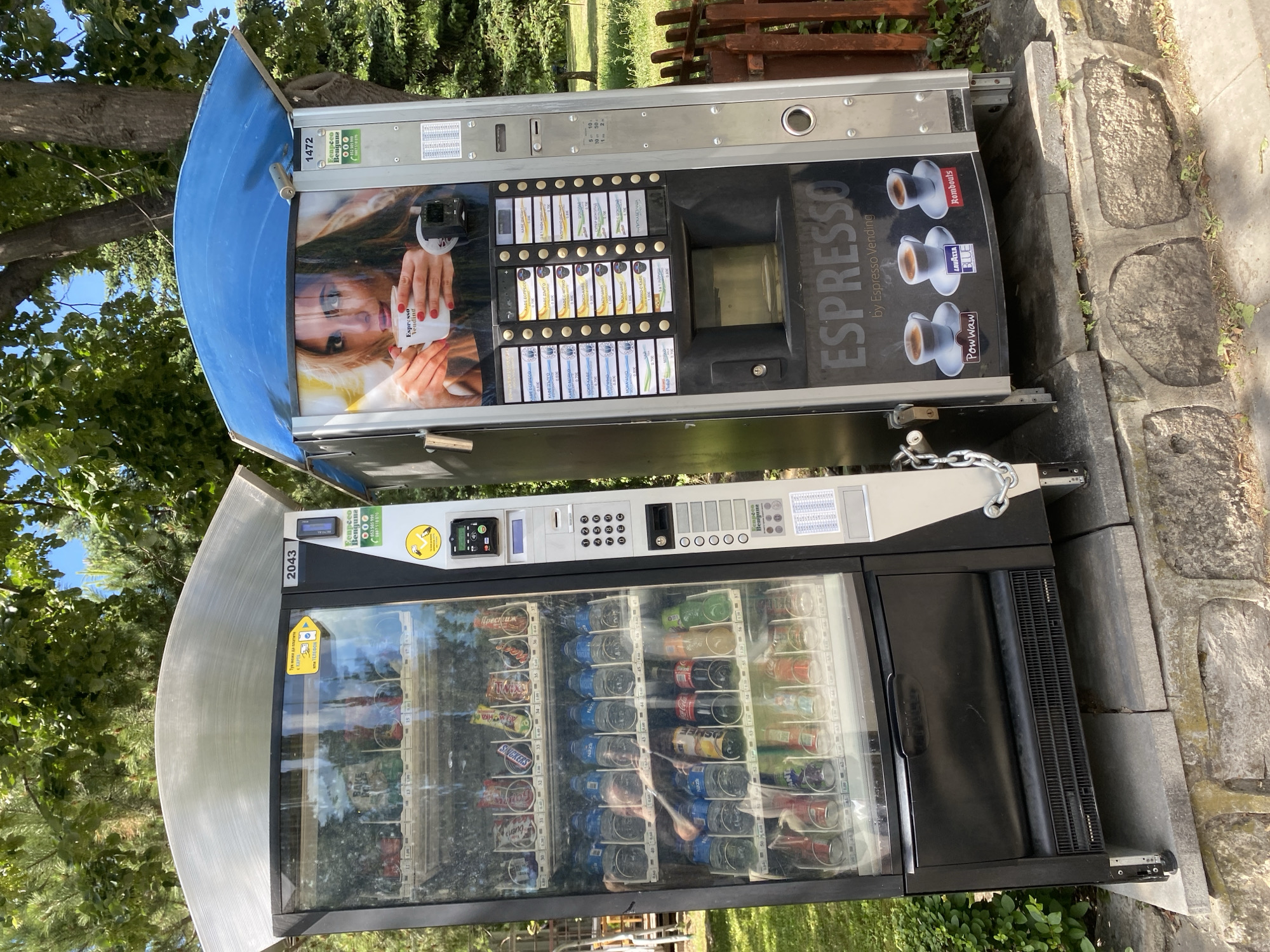 This is a forensic, matter-of-fact description of an outdoor scene in Varna, Bulgaria. Two vending machines stand side-by-side on a tiered stone base, partially obscured by a wooden fence and lush green trees. The machine on the left is a beverage vending machine filled with bottled drinks and snacks. It is silver and black with a clear glass front, displaying various products like Coca-Cola, Snickers, and Nazzi bottles. A chain and padlock secure its side. The machine on the right is a coffee vending machine with a blue canopy. It features an advertisement of a woman holding a cup of coffee. The machine dispenses various espresso-based drinks. Both machines are operational and positioned outdoors, likely in a public area. The lighting suggests it is daytime.