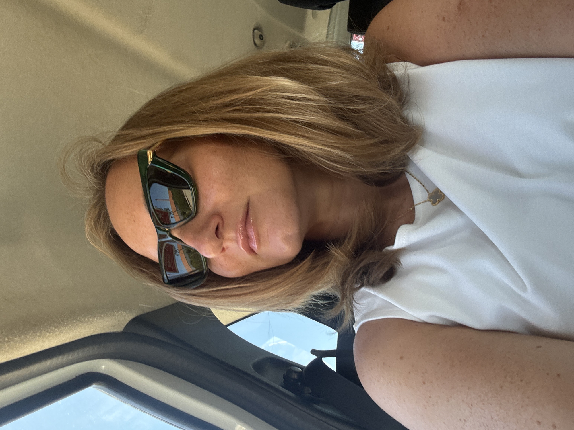 A medium shot captures a woman from the shoulders up, seated inside a vehicle. She has shoulder-length blonde hair and is wearing dark green designer sunglasses. The woman's lips are glossy, and her skin has freckles. She is wearing a white sleeveless top, and a gold pendant necklace is visible. The interior of the vehicle is visible behind her, including the grey headliner and a black seatbelt buckle. Outside the car window, the sky and some indistinct structures are visible, suggesting a daytime setting. The image appears to have been taken in Tavira, Portugal.