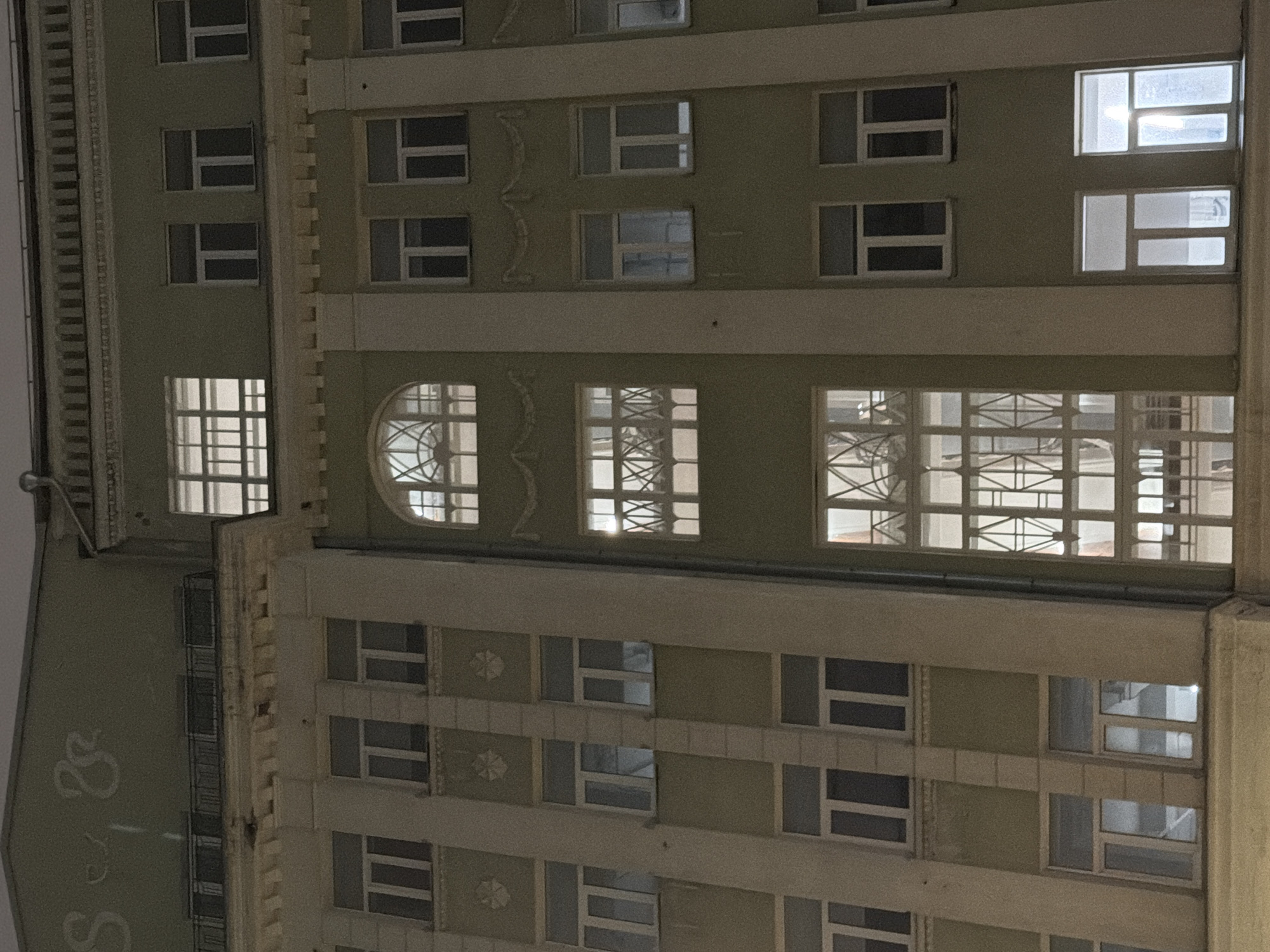 The image depicts the exterior of a building, likely an apartment complex, at night. The building is multi-storied with a facade that features numerous windows of varying sizes and shapes. Some of the windows are illuminated, suggesting occupancy. The architectural style includes decorative elements along the window frames and between floors. The overall color scheme is muted, with variations of beige and green. The photograph appears to be taken from a ground-level perspective, looking upward at a slightly skewed angle. The location is unknown.