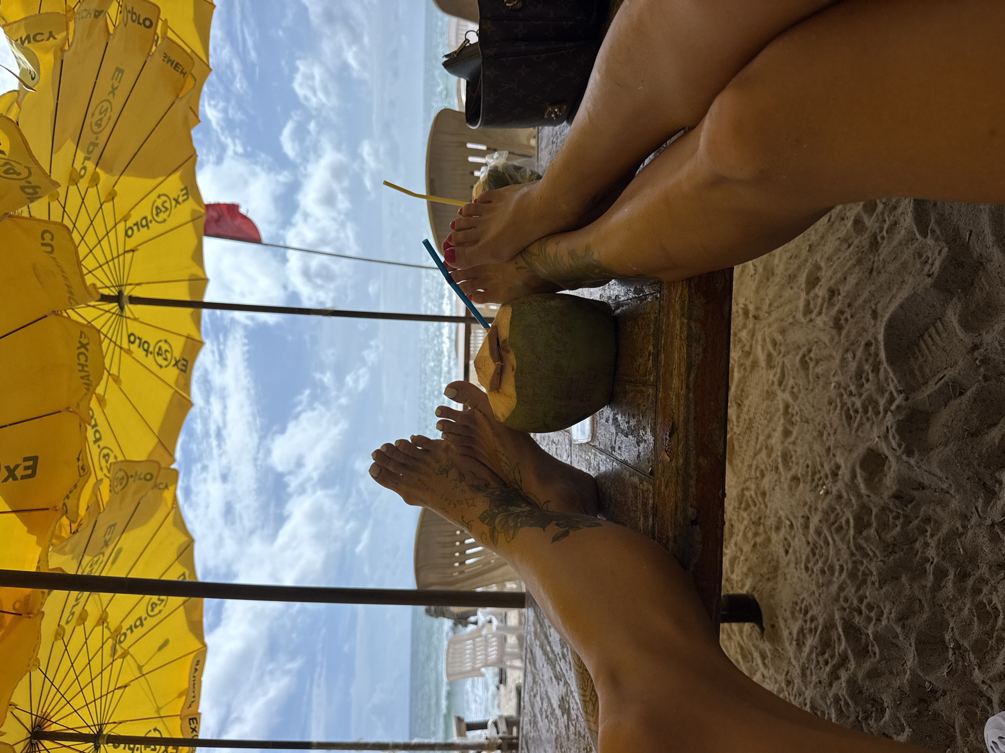 This is a medium shot, eye-level, outdoor, horizontal, daylight, close-up, point-of-view photograph taken from a beach in Choeng Thale, Thailand. The image features two pairs of bare feet resting on a wet, wooden table. Both individuals appear to have tattoos on their legs and feet.
In the foreground, a halved coconut with two straws sits on the table, suggesting a refreshing drink. To the right, a dark-colored bag is also visible on the table. In the background, the ocean stretches out under a cloudy sky, with a red flag flying on a pole. Yellow umbrellas provide shade over the scene. The beach itself is sandy and appears wet, with footprints visible. The overall impression is one of relaxation and leisure.