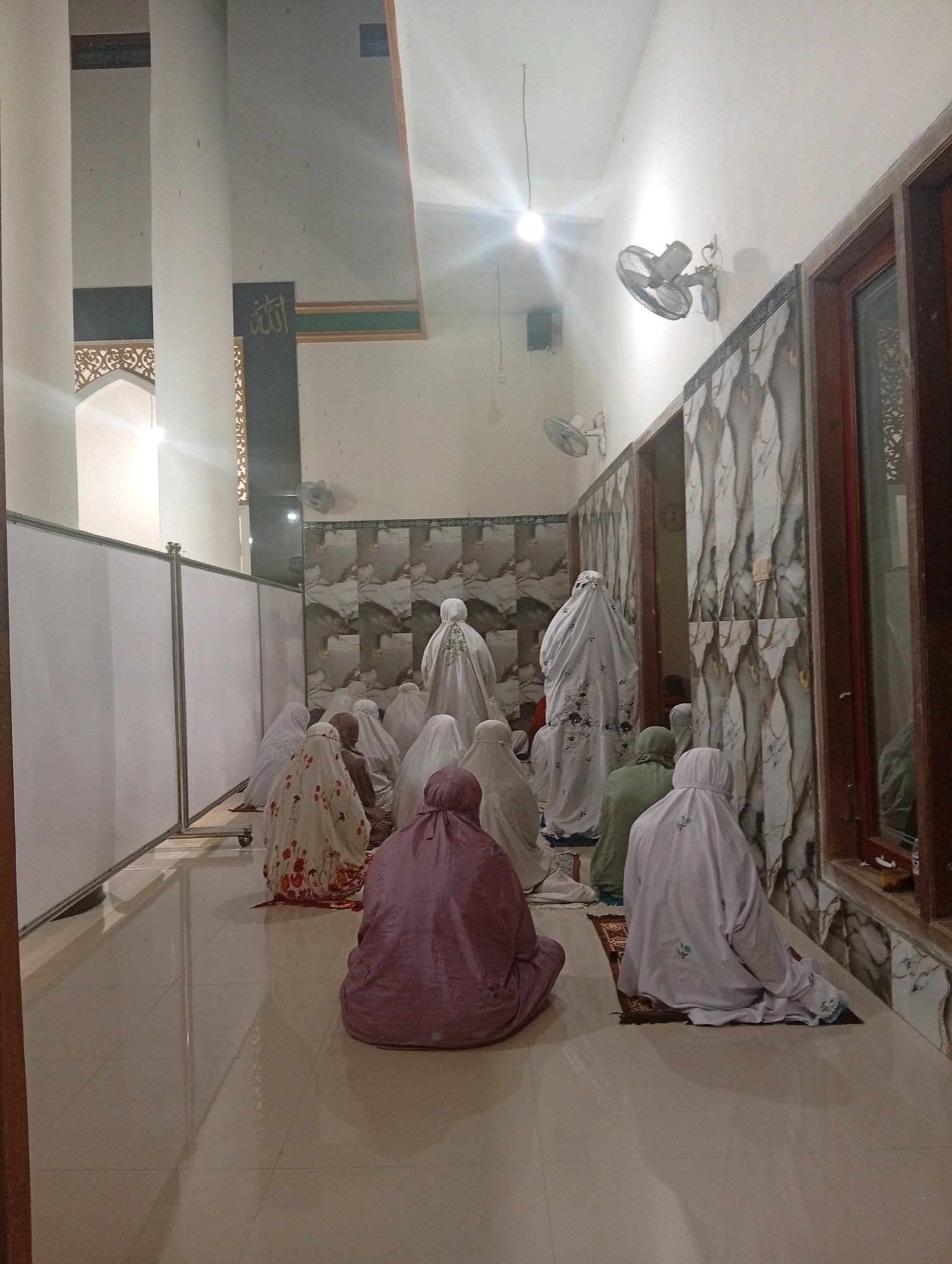 The image depicts a group of people, primarily women, praying inside a room that appears to be part of a mosque or prayer hall. They are all dressed in modest Islamic attire, covering their heads and bodies. The individuals are seated on prayer mats on a tiled floor, facing in the same direction, indicating they are performing a communal prayer. Some are in a kneeling position, while others are seated. The room has high ceilings and white walls, with decorative elements on the walls and a large window on the right side. There are also fans mounted on the walls. A partition made of white panels separates one section of the room from another. The lighting suggests it might be evening or an indoor setting with artificial lights. Based on its typical architectural style and the attire of the people, this scene is likely taking place in Trenggalek, Indonesia.