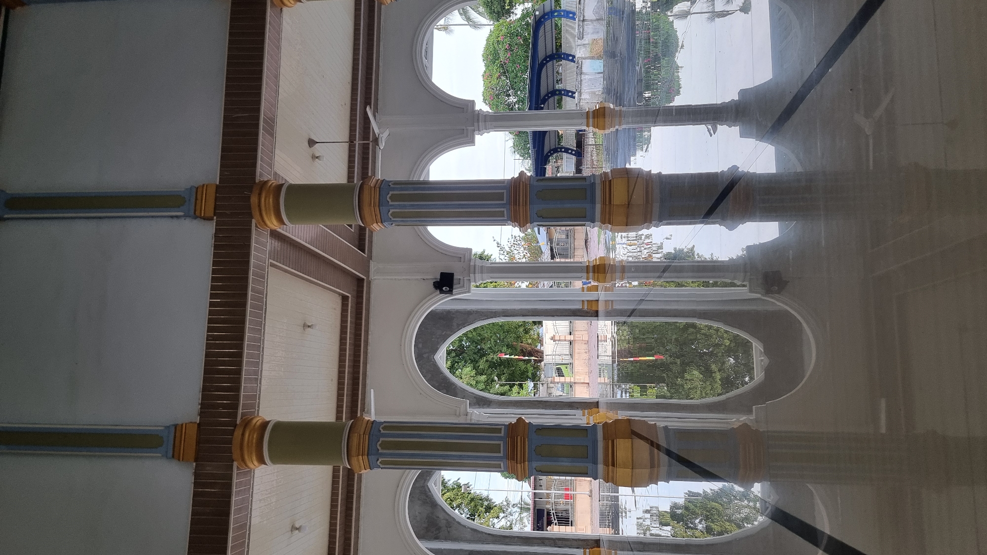 This is a vertical, eye-level photograph of a covered walkway with a highly reflective floor, likely at a mosque in Meunasahlhok, Indonesia. The walkway is supported by a series of arched openings and decorated columns. The columns are fluted and painted in alternating sections of light blue, green, and yellow. The floor is polished to a mirror-like sheen, reflecting the architecture and the greenery visible through the arches.
Through the arches, trees and a building with a blue roof can be seen. A red, white, and blue striped pole or flag is visible in the distance, near a fence. The ceiling of the walkway is made of wood panels, and there are light fixtures visible. A ceiling fan is also present in the upper right portion of the image. The overall impression is one of clean, well-maintained architecture with a strong emphasis on symmetry and reflection. There are no people visible in the image.