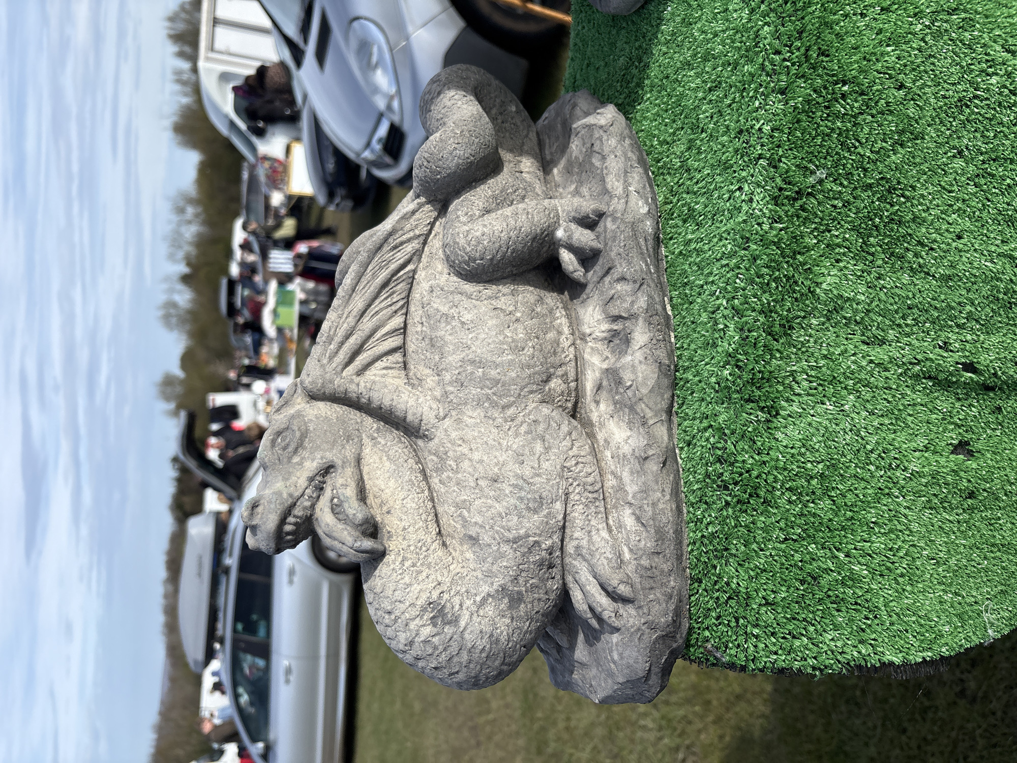 A stone gargoyle statue rests on a patch of artificial green turf. The background shows a blurred outdoor market scene with cars and people visible, suggesting a public setting. This photo was likely taken in Halton, United Kingdom.