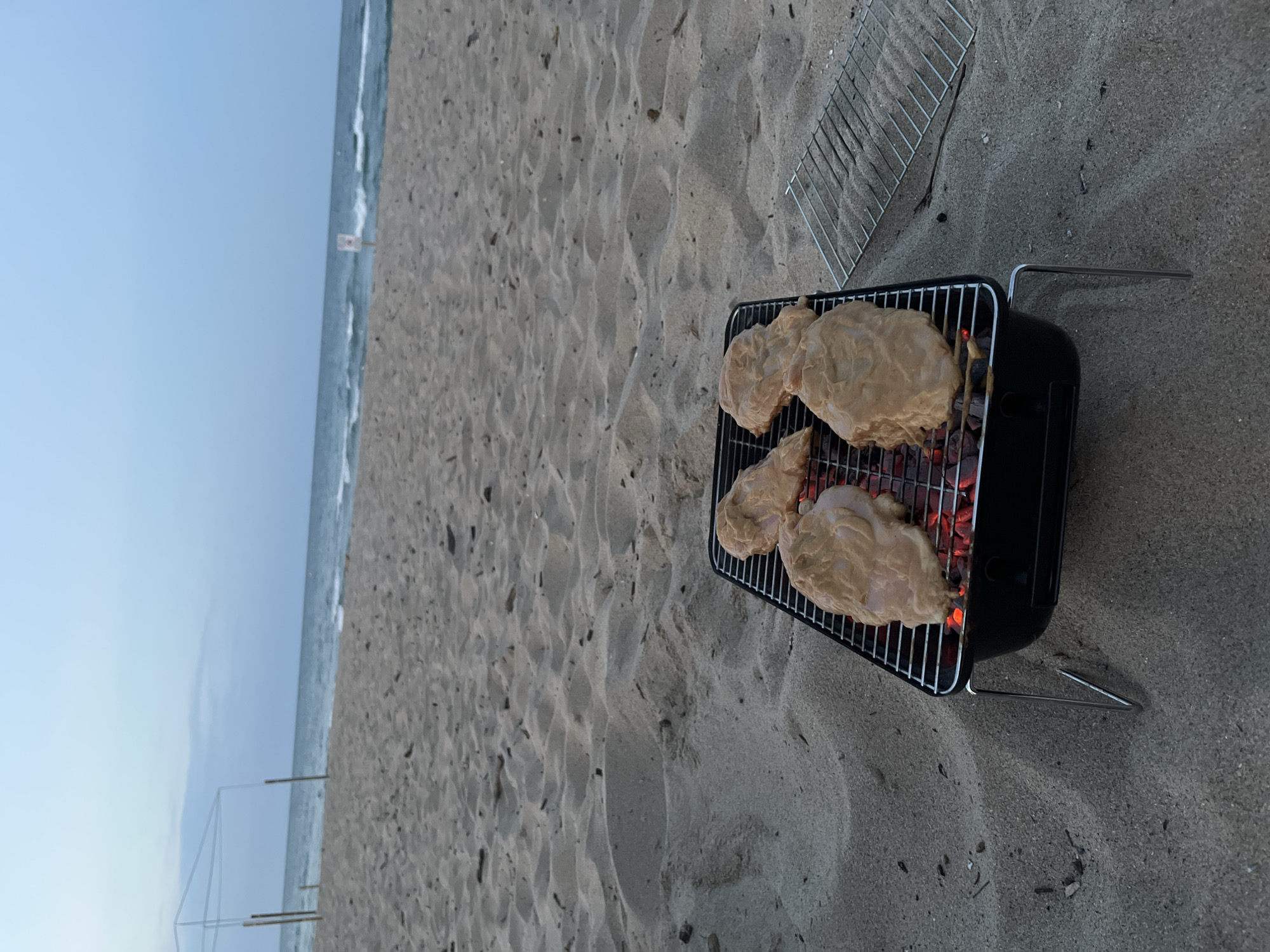 Four raw chicken breasts coated in a light-colored marinade are arranged on a black portable grill. The grill is positioned on a sandy beach. Embers are visible glowing beneath the chicken. A second, empty grill rack lies on the sand to the right of the main grill. The background consists of a wide, sandy beach with gentle waves of the sea and a pale blue sky. In the distance, near the waterline, a small white sign with red markings is visible. A partially constructed white frame structure with vertical poles is situated further down the beach on the left. The image was captured at dusk in Obzor, Bulgaria.