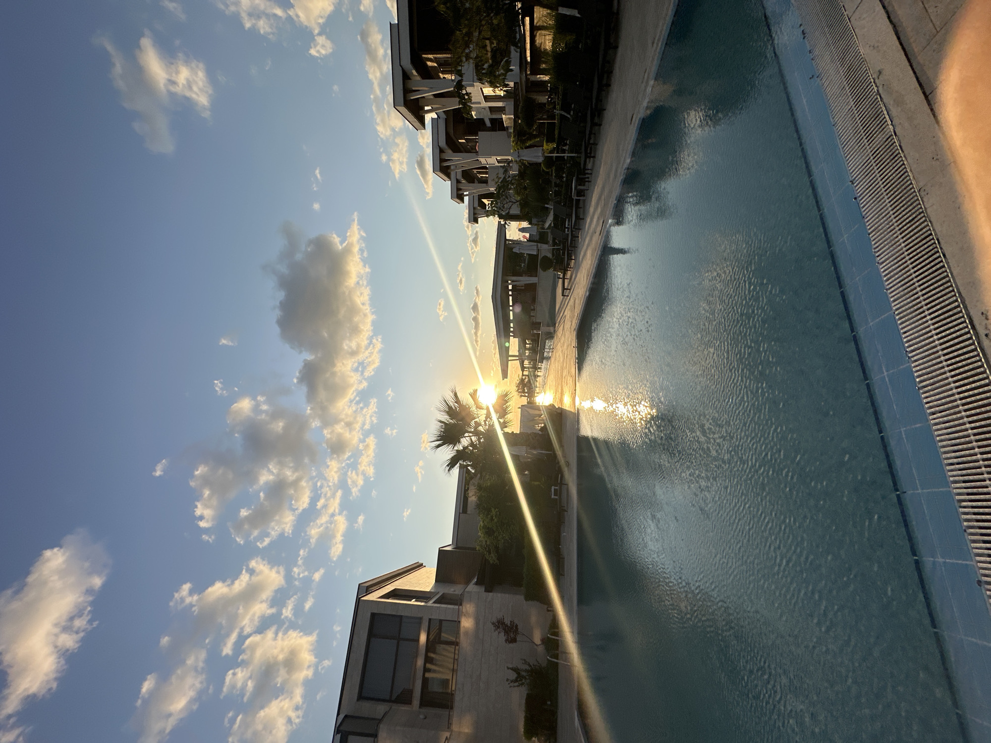 This image depicts a serene poolside scene at sunset in Sozopol, Bulgaria. The foreground features a clear blue swimming pool with rippling water, reflecting the golden hues of the setting sun. The pool is bordered by a tiled edge and a metal grate drain. In the background, modern architectural buildings with clean lines and flat roofs are visible, interspersed with palm trees and lush greenery. The sky is a vibrant blue, dotted with fluffy white and golden clouds illuminated by the sun's rays, which create a strong lens flare effect across the scene. The overall impression is one of tranquility and luxury, characteristic of a resort setting.