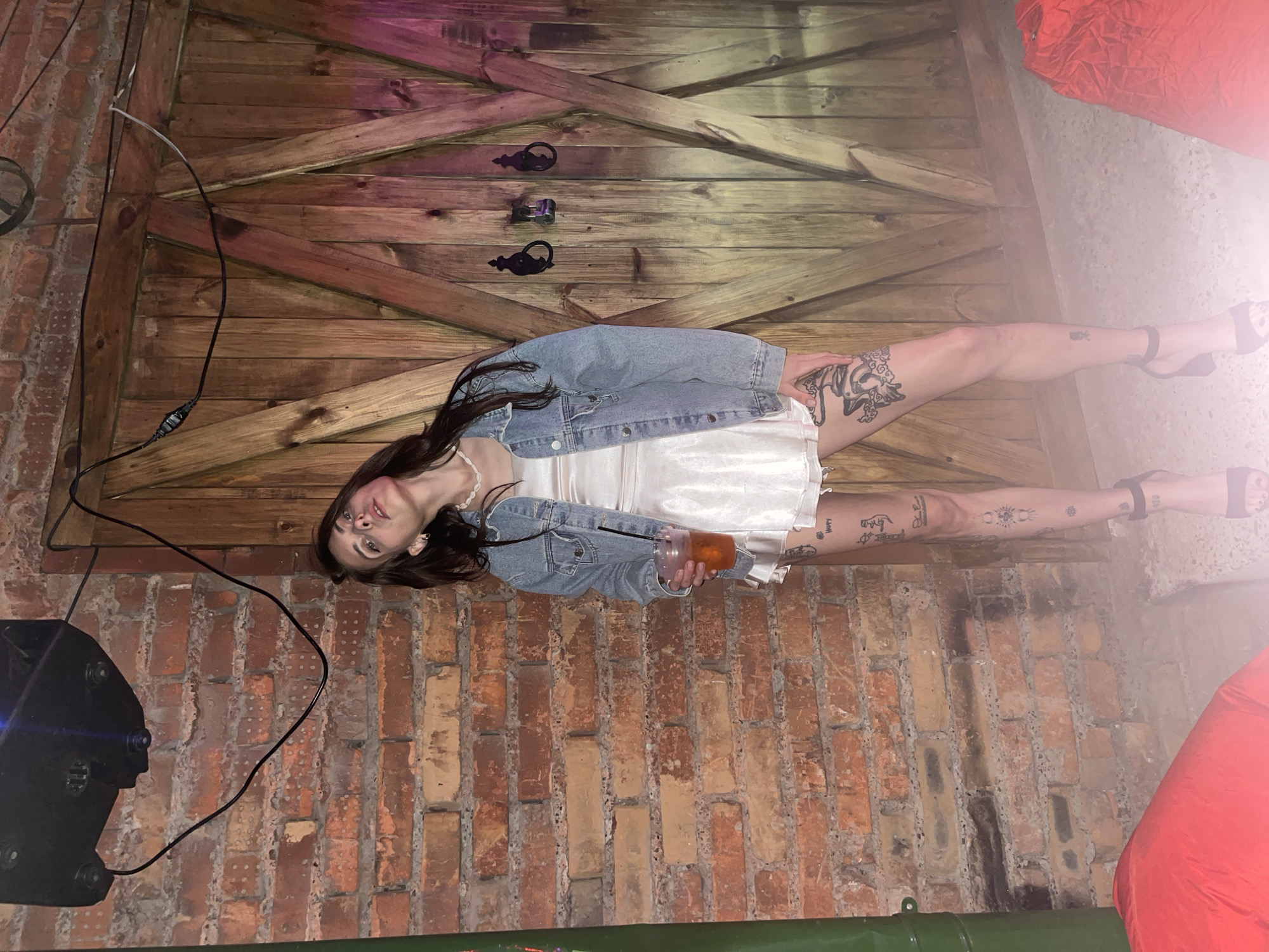 A woman stands in front of a wooden door, holding a drink. She is wearing a denim jacket over a white dress and black heels. She has tattoos on her legs. The background appears to be a brick wall and a wooden door. There is a light fixture above her and some red objects on the floor in the foreground. The image was taken in Krasnoyarsk, Russia.