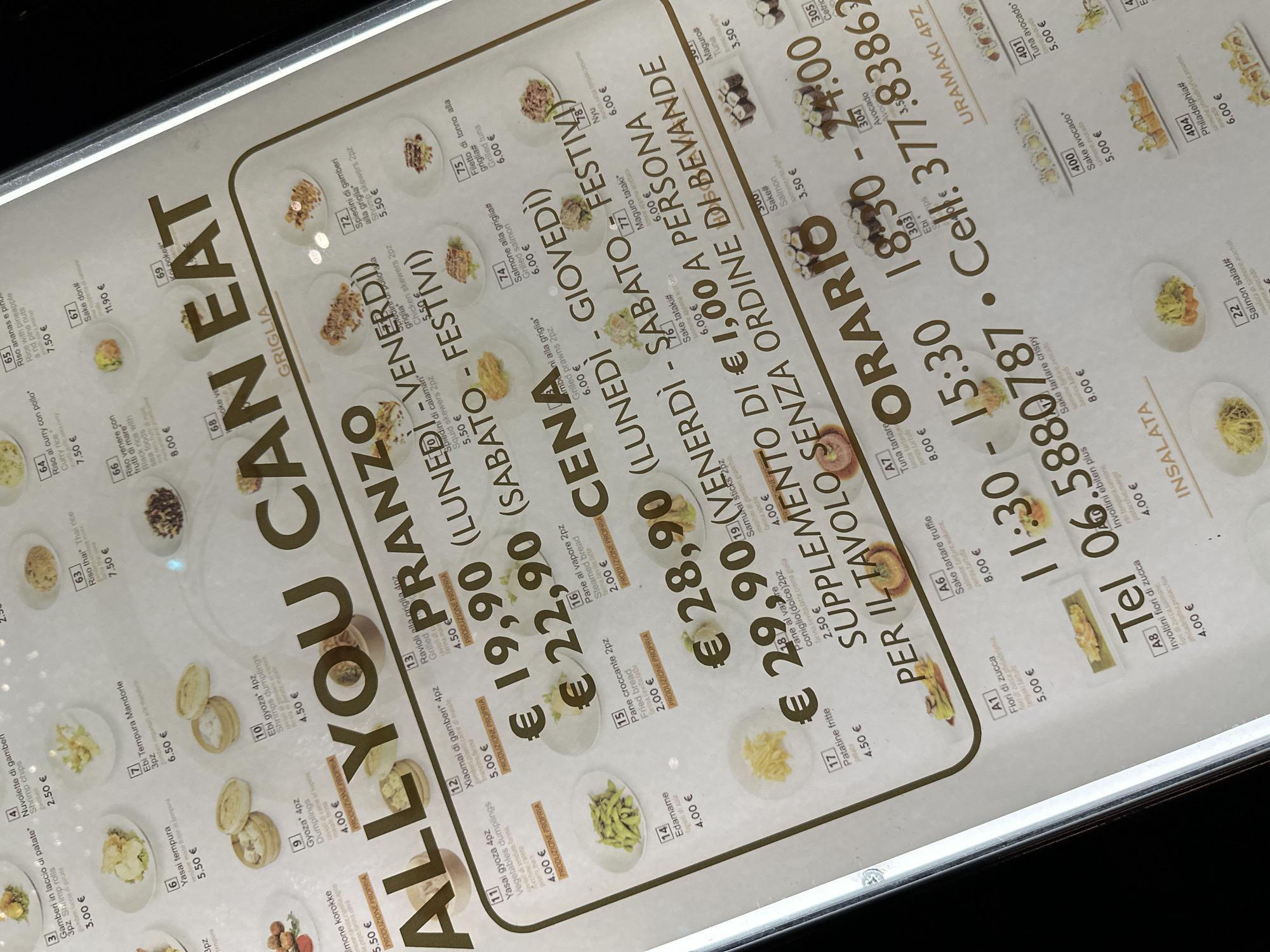 The image displays a close-up of a menu, likely from an all-you-can-eat Japanese restaurant, featuring a variety of sushi and other dishes. The menu is presented in Italian and English, with prices listed in Euros. Many items are accompanied by small photographic samples. The prominent text "ALL YOU CAN EAT" suggests the restaurant's dining concept. Below this, there are sections for "PRANZO" (Lunch) and "CENA" (Dinner), with daily breakdowns for weekdays and weekends. Further down, there are details about supplementary charges and opening hours, including a phone number and a cell number. The overall impression is that of a well-organized and appealing menu designed to attract customers to a Japanese dining experience. There are no people visible in the image.