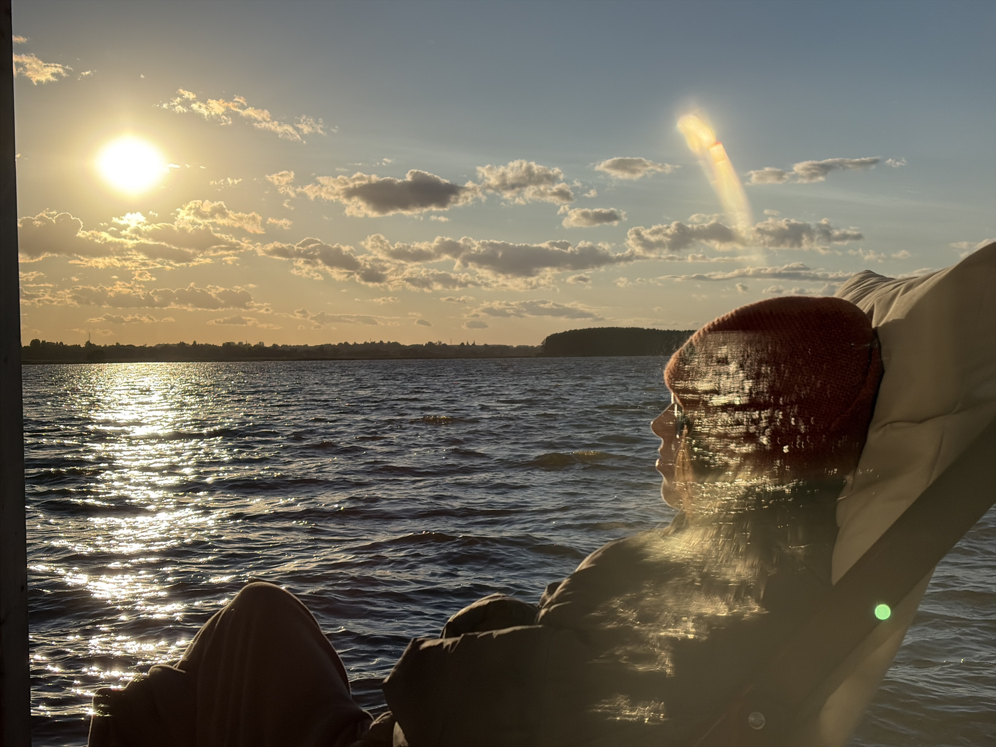 A side profile of a person wearing a red knit hat and a dark jacket is captured as they relax in a chair. They are looking out at a body of water, which is shimmering with sunlight. The sun is low in the sky, casting a warm glow and illuminating a trail of light on the water's surface. In the distance, a shoreline with trees and a faint silhouette of buildings can be seen. The sky above is a mix of blue and scattered clouds, with a peculiar lens flare creating a vertical, bright streak. The image appears to have been taken in Vidogoshchi, Russia, during a sunset.