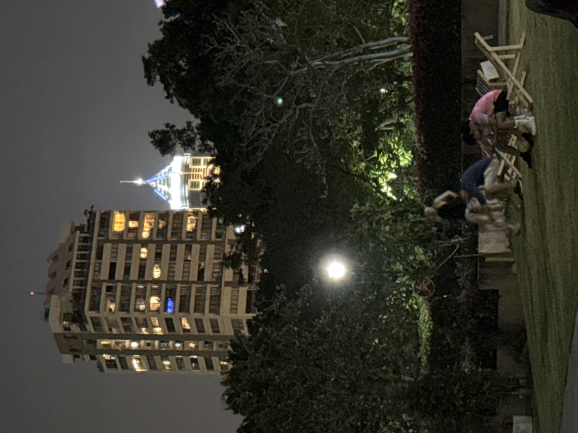 At night, a tall building in Bangkok, Thailand, is viewed from a park. The building is lit with yellow lights, with a spire on top illuminated in white and blue. A bright streetlamp shines near a collection of trees and bushes. In the park, people sit on chairs, engaging in conversation. The scene is typical of an urban evening.