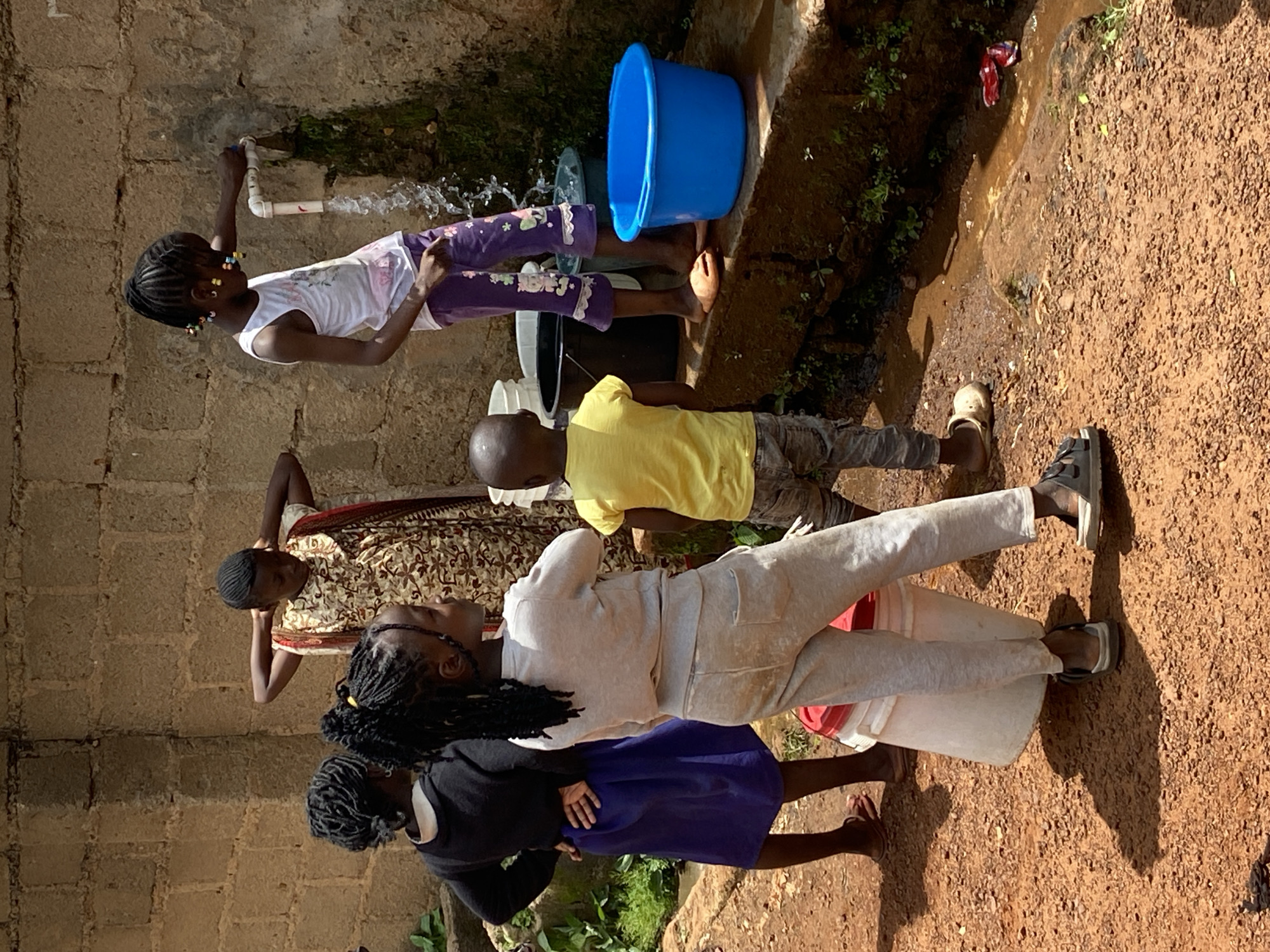 In the city of Bukuru, Nigeria, a group of five children gathers around an outdoor water tap. The scene depicts a brick wall as a backdrop and a reddish-brown, gravelly ground. The children, varying in age, are engaged in activities related to water collection.
The child on the far right, a girl wearing a white tank top and purple patterned pants, is actively turning on the tap, causing water to stream down the wall and into a collection basin. She has braids adorned with colorful beads. Next to her, a younger boy in a yellow t-shirt and grey pants stands observing the water flow.
To the left, a girl in a black long-sleeved shirt and a purple skirt is facing away from the tap, with her hands on her hips. Beside her, another girl, also facing away, wears a light grey sweatshirt and matching sweatpants. She is carrying a large white bucket with a red lid. The eldest child, standing further back and partially obscured, wears a patterned dress and has her hands clasped behind her head.
Several containers are strategically placed to collect the water: a bright blue basin is directly below the tap, and other buckets are visible around the area, including the white one being carried. The environment appears to be a utilitarian outdoor space, possibly a courtyard or a communal area, where water access is facilitated by the tap and collection containers. The ground is uneven and composed of small stones and dirt, with some sparse vegetation growing along the base of the wall.