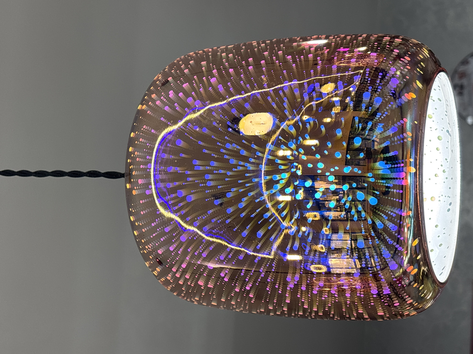 A glass pendant lamp, viewed from below, dominates the frame. The lamp has a bronze-colored, transparent body with small, evenly spaced dots across the surface, creating a textured effect. Inside the glass, patterns of blue and purple light emanate outward, seemingly reflecting off internal surfaces. A distinct brighter yellow circle appears within the center. The lamp is suspended by a black cord attached to the left. The right-hand side of the lamp appears to be open, with a white, reflective interior. The background is a flat, light gray, indicative of a wall or ceiling. The location in Gorna Malina, Bulgaria is not discernible from the image.