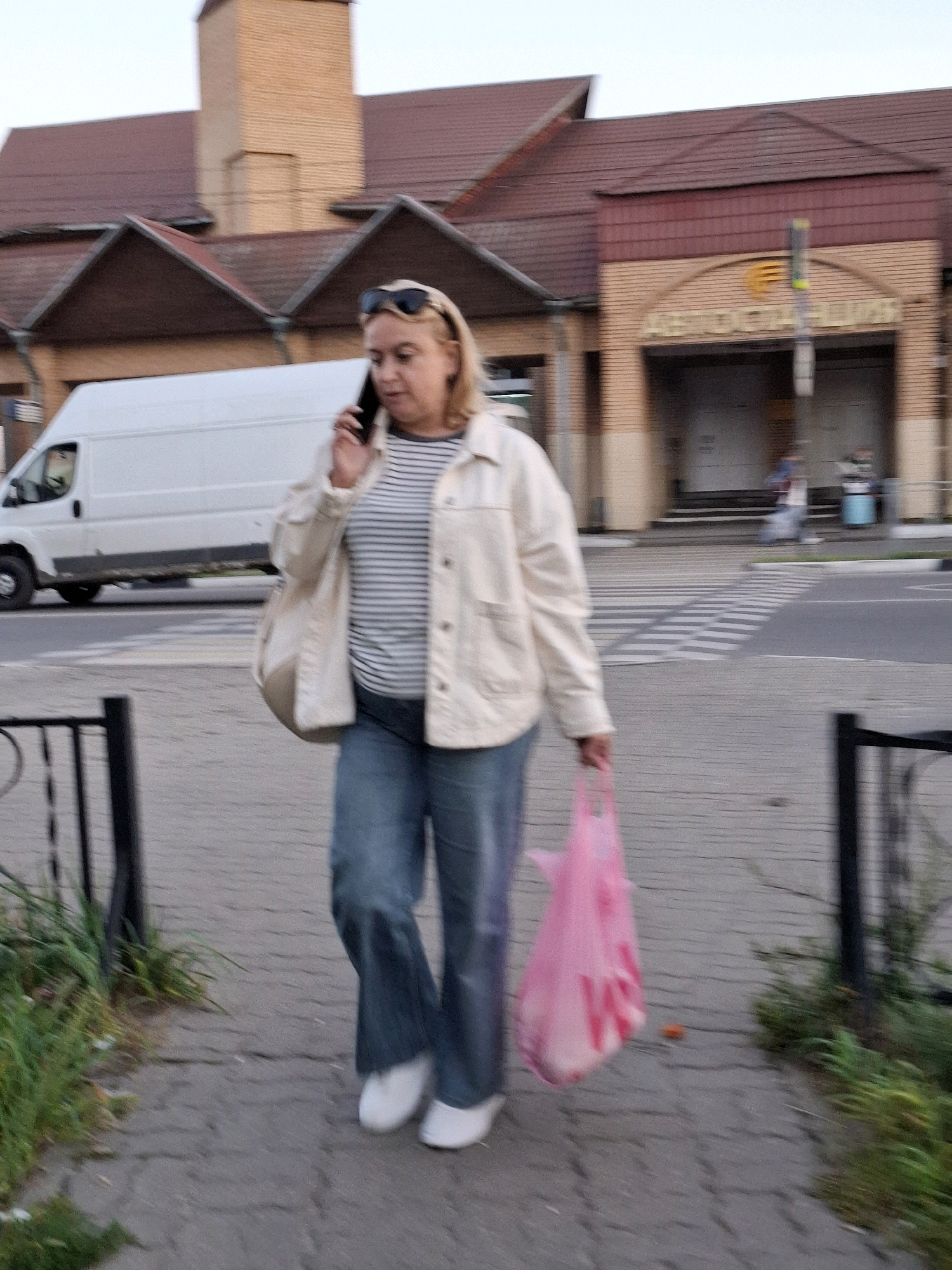 A woman with blonde hair is walking on a sidewalk and talking on her cell phone. She is wearing a cream-colored jacket, a striped t-shirt, and blue jeans. She is carrying a pink plastic bag in her right hand and a purse over her left shoulder. A white van is parked behind her on the street. The background shows a brick building with a red roof. The scene appears to be an outdoor shopping area.