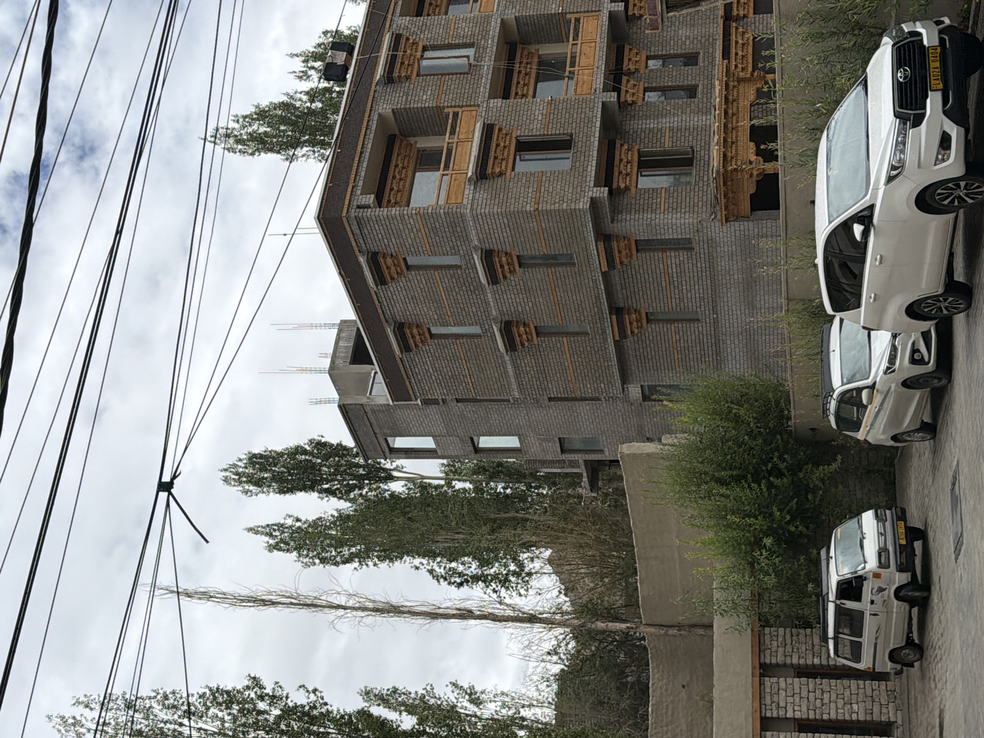 A side view shows a modern, multi-story building constructed with grey bricks, featuring ornate wooden window frames and balconies with intricate carvings. The building is located in the city of Leh, India, as indicated by the license plates on the vehicles. In the foreground, two white cars are parked on a paved surface. The car on the left is a white van with a roof rack. The car on the right is a white SUV. Tall, slender trees with green foliage line the side of the building, and a grey concrete wall with a textured finish runs along the foreground. Power lines are visible against a cloudy sky.