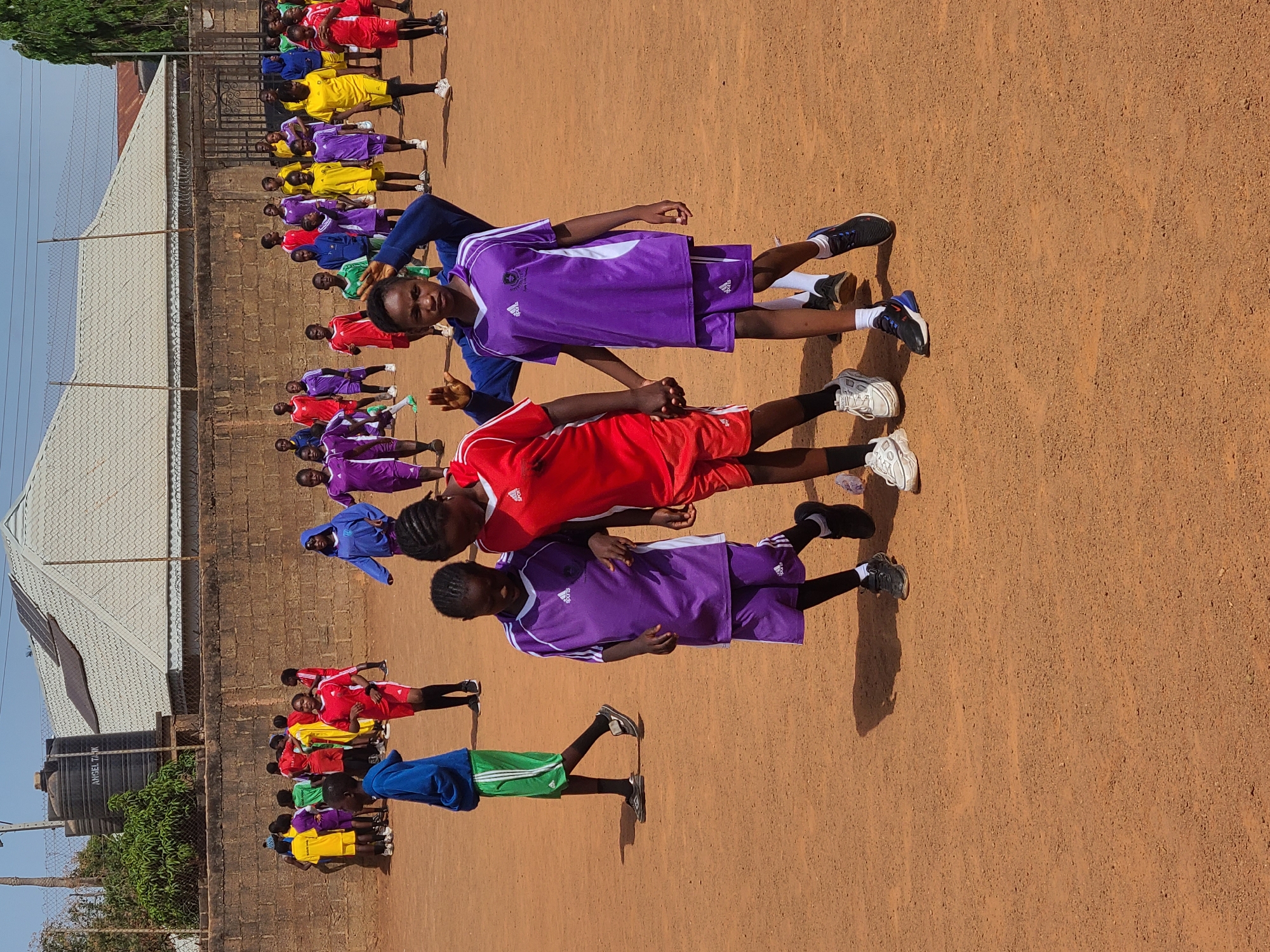 The image shows a group of children standing in a line in Barakin Lerawi, Nigeria. They are wearing brightly colored athletic uniforms in hues of red, purple, yellow, green, and blue. The children are standing on a dirt surface. A building, fence, and water tank are visible in the background.