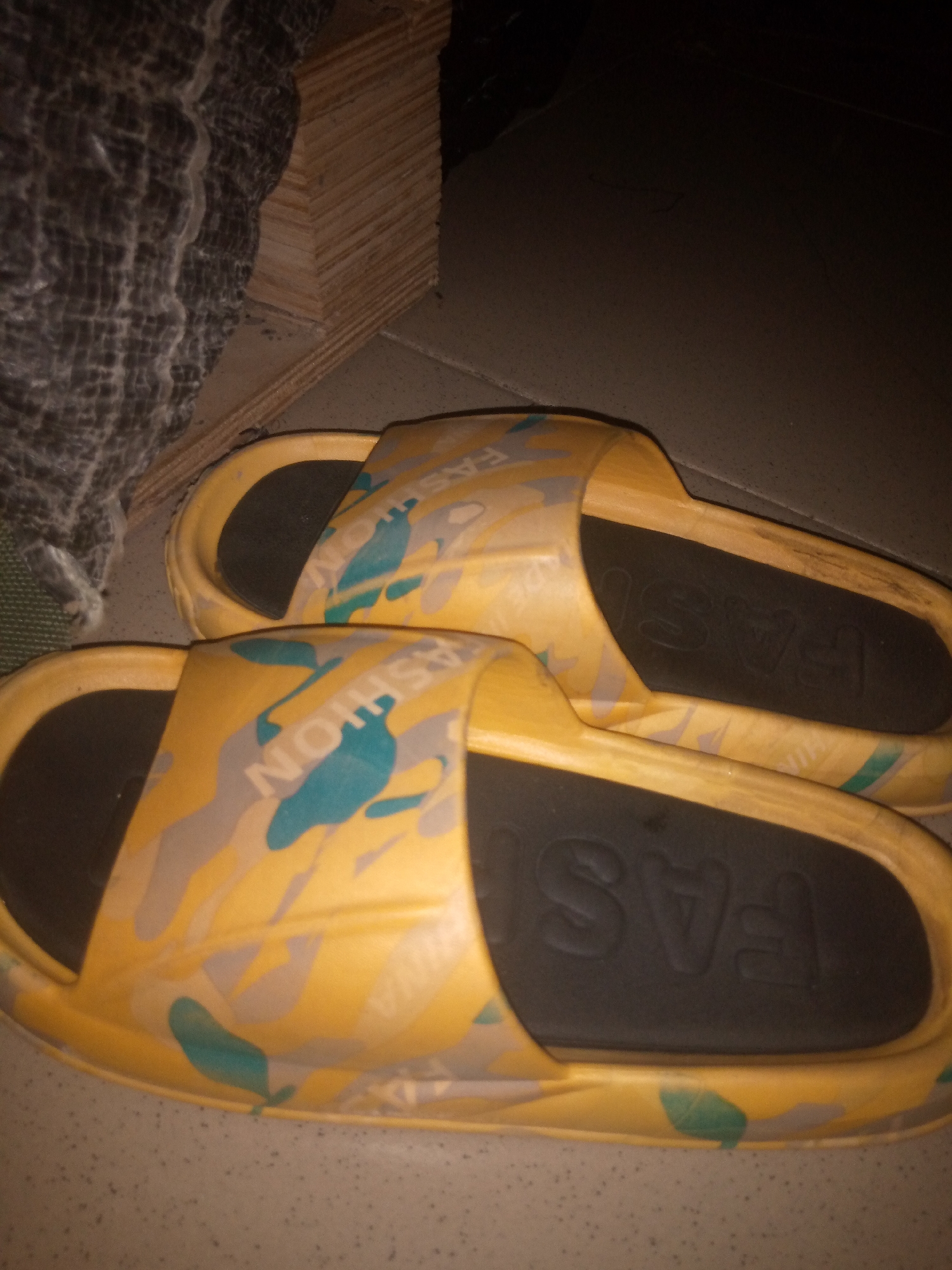 Two yellow slides with a camouflage pattern of gray and teal are displayed on a speckled gray floor. The word "FASHION" is embossed on the black insole of the slides. To the left of the slides is a textured, dark gray material. Behind the slides, a stack of light brown wooden planks is visible, leaning against a dark surface. The lighting suggests an indoor environment with low ambient light.