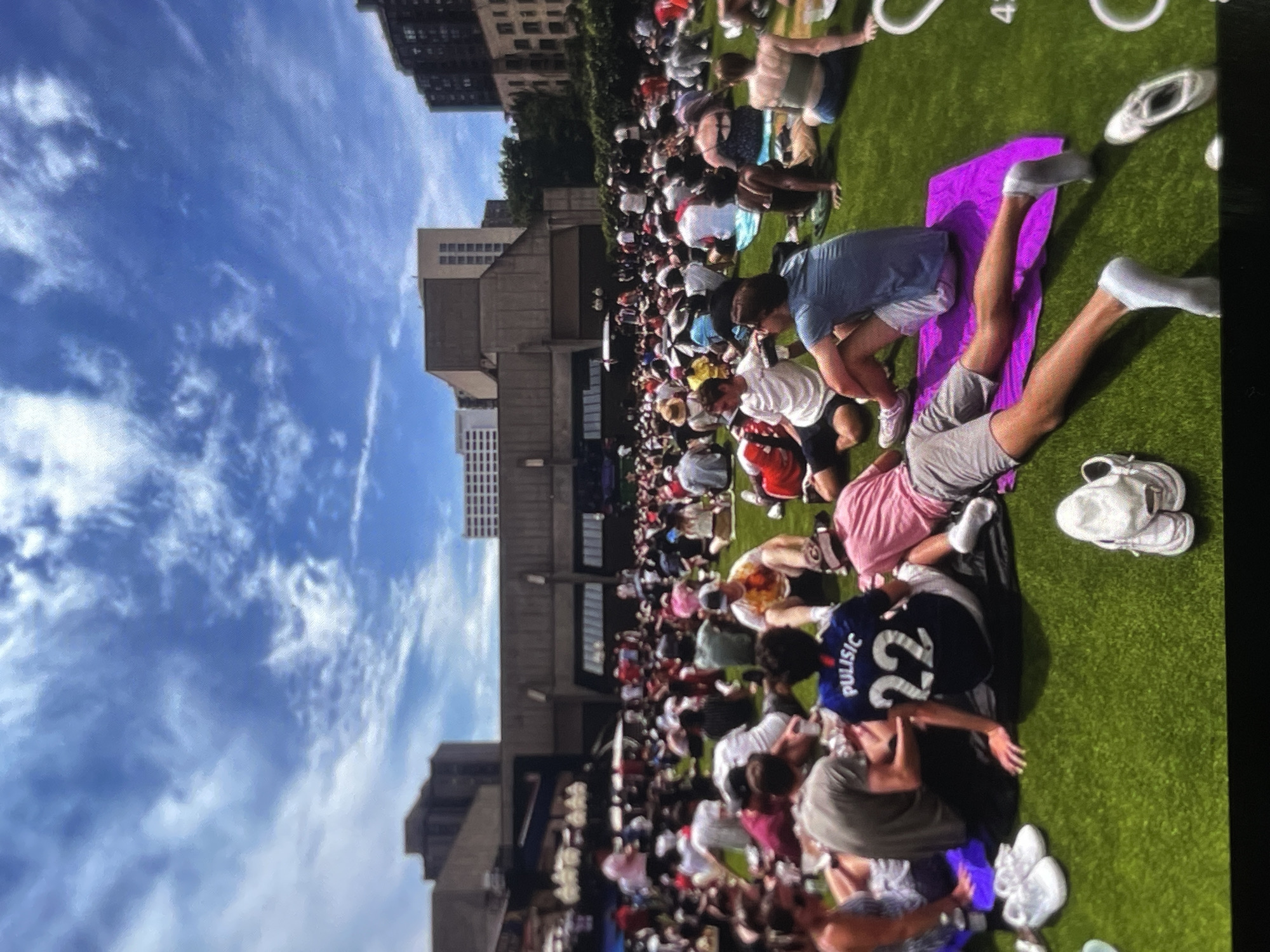 A crowd of people are sitting on a grassy area in front of a building. The sky is blue with scattered clouds. A person in the foreground is wearing a blue soccer jersey with "Pulisic" and the number "22" on the back. Another person is wearing a pink shirt and grey shorts. Several pairs of white sneakers are visible on the grass. In the background, there are several tall buildings. The scene appears to be an outdoor gathering or event.