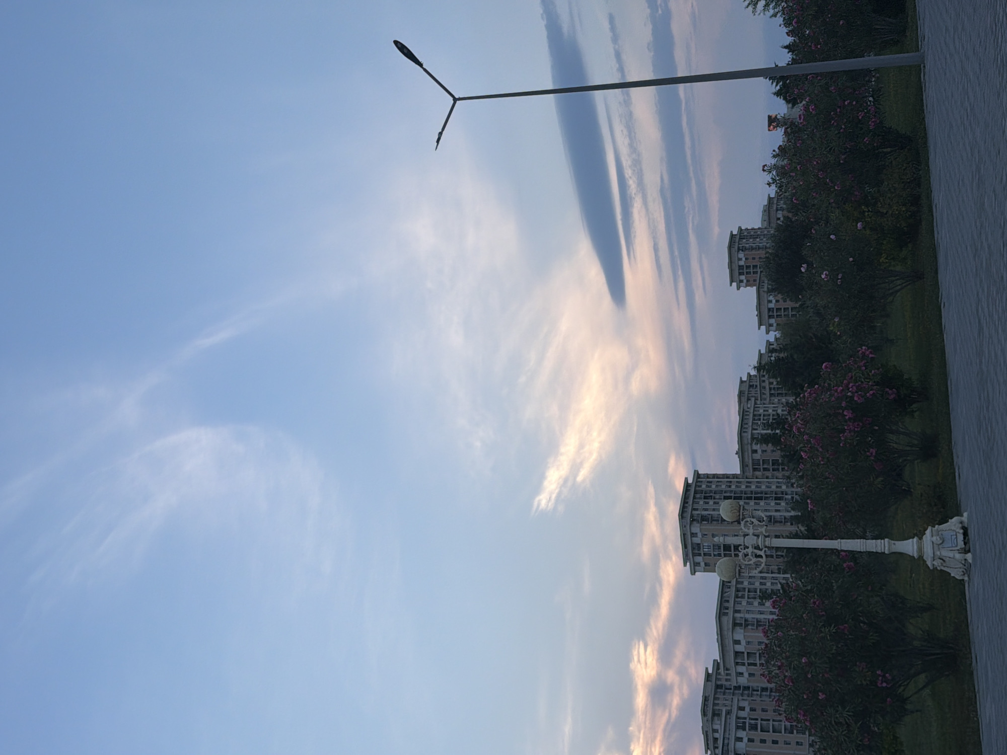 The image depicts a view of apartment buildings in Sumgayit, Azerbaijan, on a cloudy day. The sky is a mix of blue and pale pink hues, with wisps of clouds stretching across it. In the foreground, there is a white lamppost with ornate detailing. Behind the lamppost, there are bushes with pink flowers, and behind those, several multi-story apartment buildings are visible. A modern streetlamp stands to the right of the frame, taller than the ornate lamppost. The ground in the foreground is paved with gray interlocking bricks. The overall impression is that of an urban park or boulevard setting at dusk or dawn.