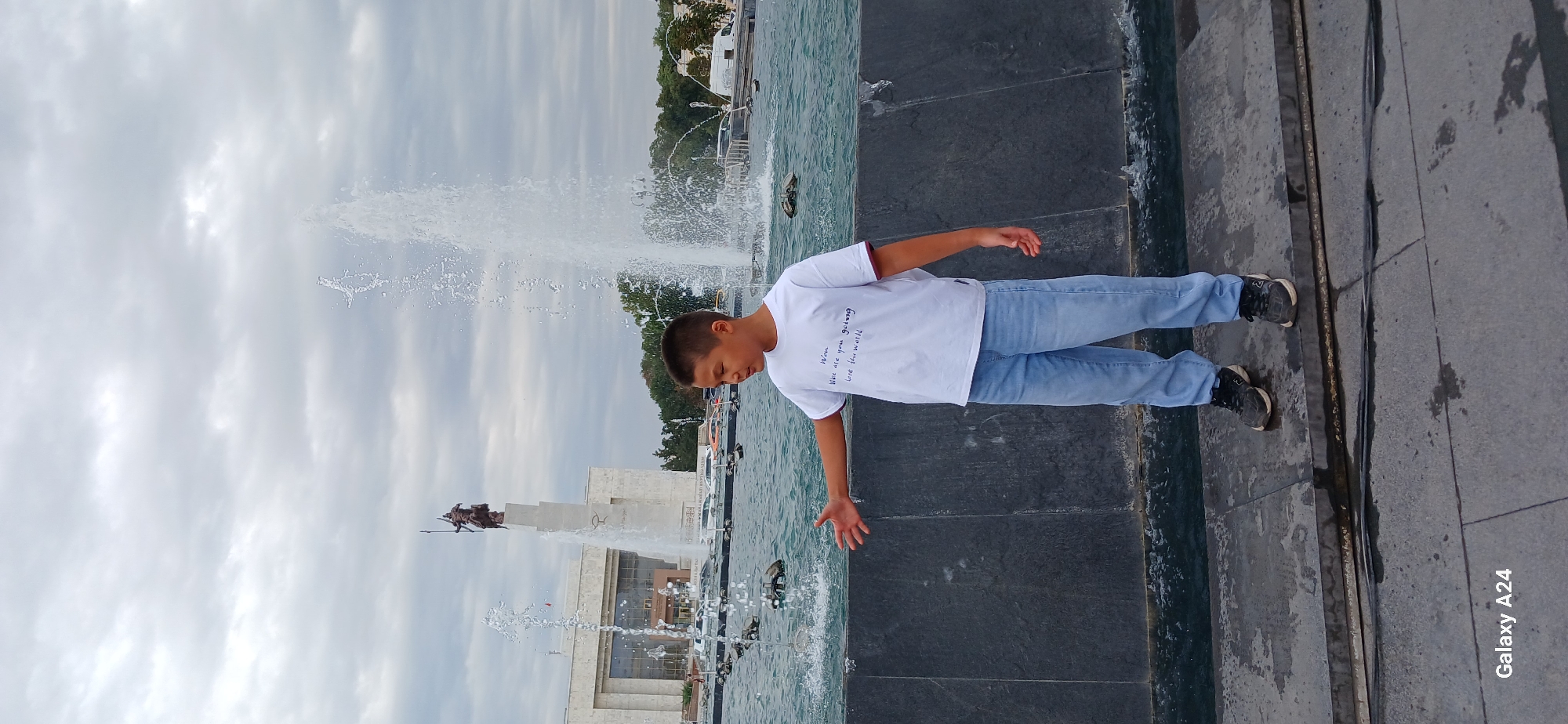 A young boy stands in front of a large fountain in Bishkek, Kyrgyzstan, with a statue and building in the background. The boy, wearing a white t-shirt with writing on it and light blue jeans, faces the fountain with his left arm outstretched towards the water. The fountain features multiple jets of water, some reaching high into the sky. Behind the fountain, a grand building with columns is visible, and atop a tall pedestal, a statue of a warrior on horseback stands against a cloudy sky. The area around the fountain appears to be paved with dark stone. The overall scene suggests an outdoor public space, likely a park or square in the city.