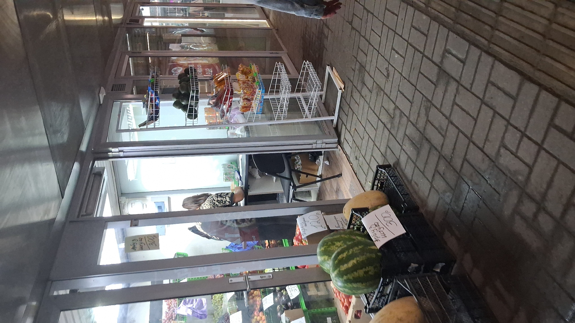 The image depicts a fruit and vegetable stall, likely in an outdoor market, with a covered ceiling and glass-fronted stalls. The ground is paved with interlocking bricks, some of which appear wet. In the foreground, several watermelons and other produce are displayed in black plastic crates, with price tags visible. One price tag reads "арбуз штучно 300р" (watermelon, per piece, 300 rubles). Another price tag in the background indicates "120р".
Inside one of the stalls, a woman with long brown hair, wearing a leopard-print top and black pants, is seen behind a counter. She appears to be interacting with a customer or handling transactions. Various fruits and vegetables are visible in crates and on shelves within the stalls. A metal shelving unit displaying packaged snacks and produce like avocados is also present. The overall lighting suggests it is either evening or an overcast day. The presence of Cyrillic script on the price tags and the style of the market suggest the location is likely in Russia, possibly Novosibirsk, given the context provided.