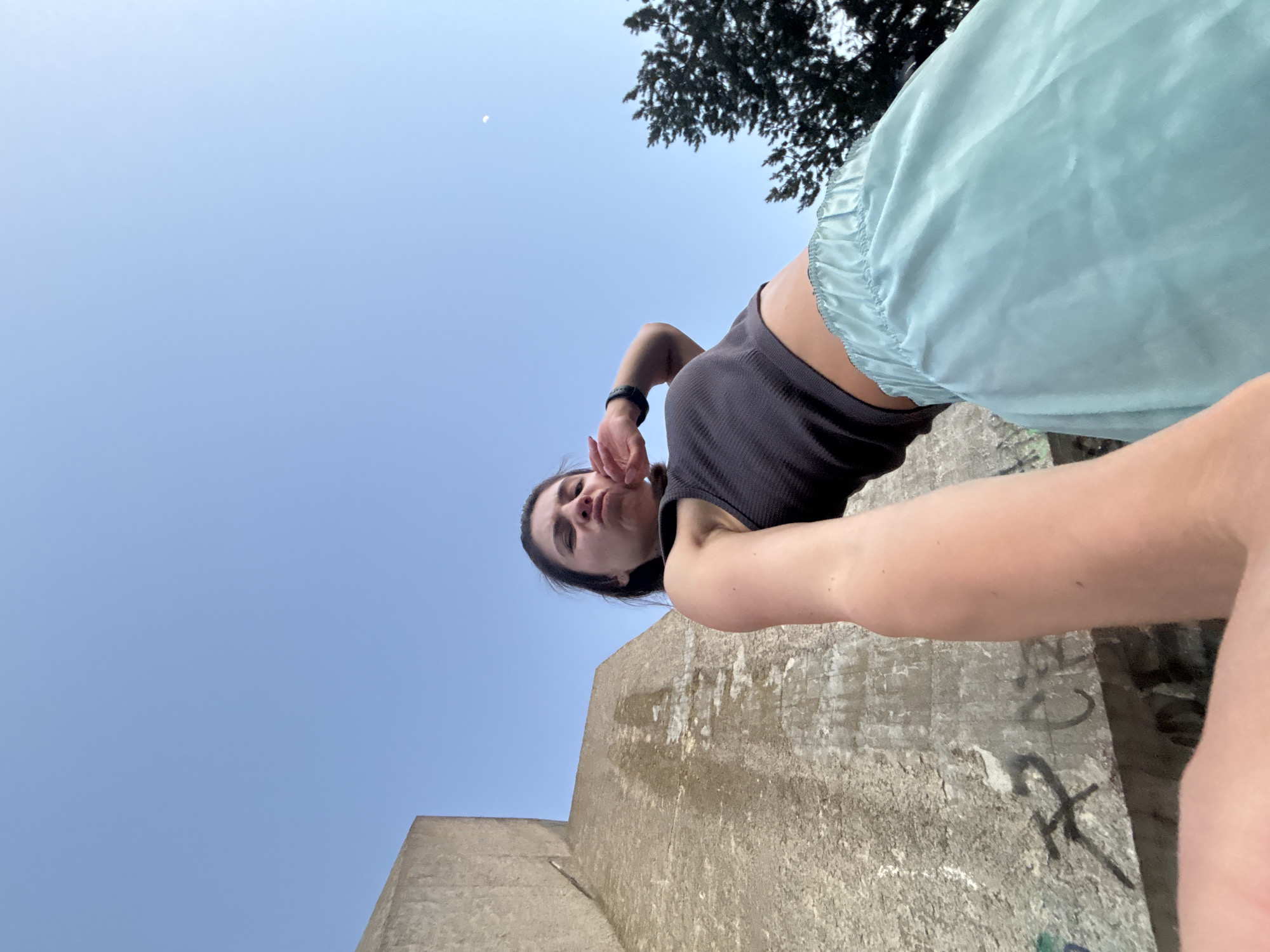 A low-angle, wide shot shows a young woman posing in front of a Brutalist concrete structure. The sky above is clear and blue, with a sliver of a crescent moon visible. The woman, wearing a dark grey tank top and light blue satin shorts, pouts with her hand near her mouth. Graffiti is present on the concrete structure. The scene is outdoors, with a tree visible in the background. The concrete structure appears to be part of a historical monument or a building with a distinct architectural style. The overall impression is a snapshot taken during the day, possibly in an urban park or historical site.