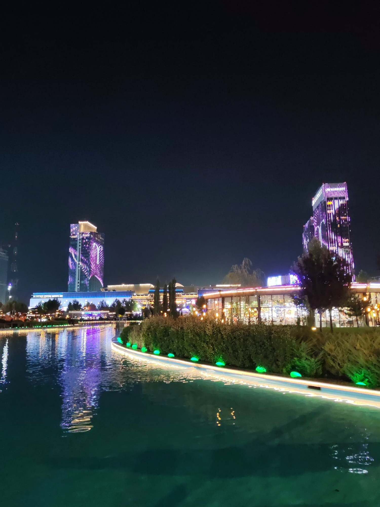 This image captures a nighttime scene at the Tashkent City Congress Hall and residential complex in Tashkent, Uzbekistan. A body of water in the foreground reflects the colorful lights of the surrounding buildings and the night sky. Along the edge of the water, there is a pathway lined with green lights and lush green bushes. In the background, two prominent modern buildings are illuminated with purple and pink geometric light patterns. The building on the right, a hotel, displays the "Marriott" logo in illuminated letters near its top. A lower building with a glass facade, likely part of the commercial or entertainment area, is also visible, adorned with numerous warm-toned lights. Trees are scattered throughout the scene, some bare and others with foliage, adding to the urban landscape. The overall ambiance is that of a vibrant and illuminated modern city center at night.
