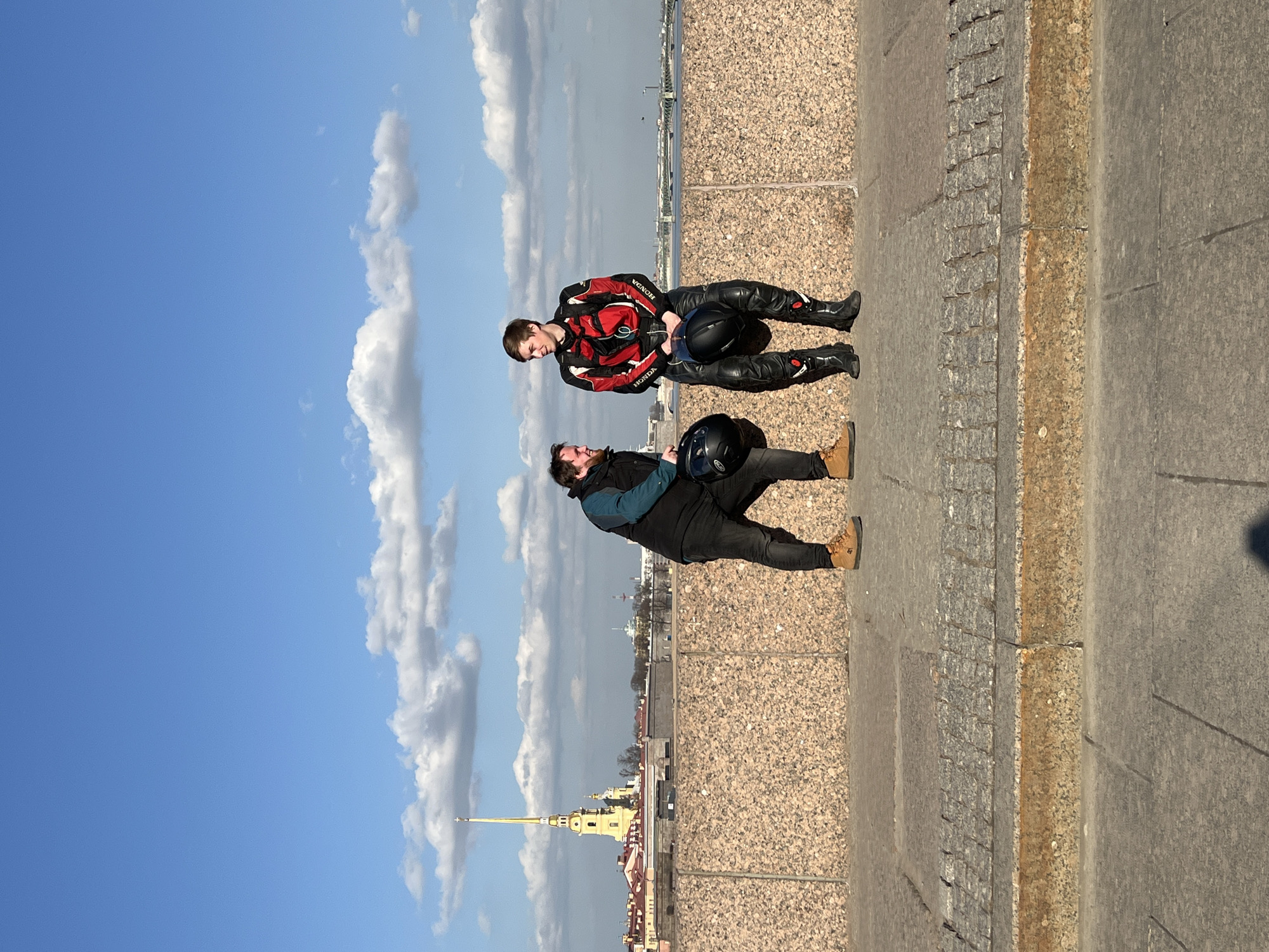 The scene captures two adult males standing on a speckled granite embankment in Saint Petersburg, Russia, under a blue sky with scattered white clouds. The subject on the left, an adult male with a beard, is dressed in a dark vest, a dark teal long-sleeved shirt, dark pants, and tan work boots. He holds a black motorcycle helmet in his left hand and is observed looking towards the second subject. The subject on the right, a younger adult male, is clad in a red and black leather motorcycle jacket with 'HONDA' lettering, matching leather pants, and black motorcycle boots. He holds a black motorcycle helmet in front of him, between his knees, and is also observed looking towards the first subject. The background features the prominent golden spire of the Peter and Paul Cathedral, situated within the Peter and Paul Fortress. Beyond the fortress, the Neva River is visible, extending to the right with a distant bridge. The ground in the foreground is paved with large grey flagstones, bordered by a strip of darker cobblestones adjacent to the granite embankment wall.