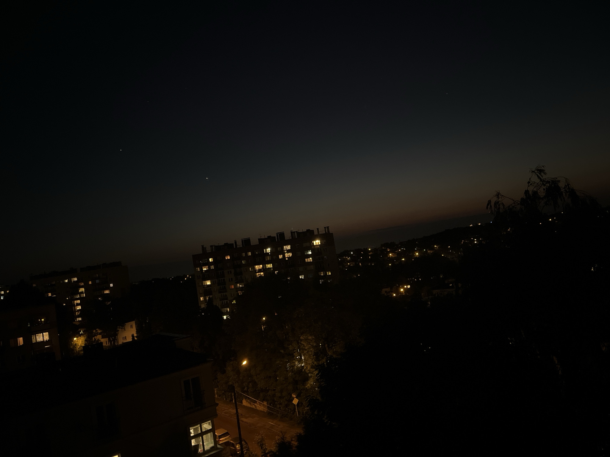 The image depicts a nighttime urban scene in an unknown city. Several apartment buildings are visible, some with illuminated windows, suggesting residential occupancy. Streetlights cast a warm glow on the roads and surrounding vegetation, which includes trees and bushes. The sky is dark with a slight gradient towards the horizon, indicating the presence of residual twilight or light pollution. A few stars are faintly visible in the upper portion of the image.