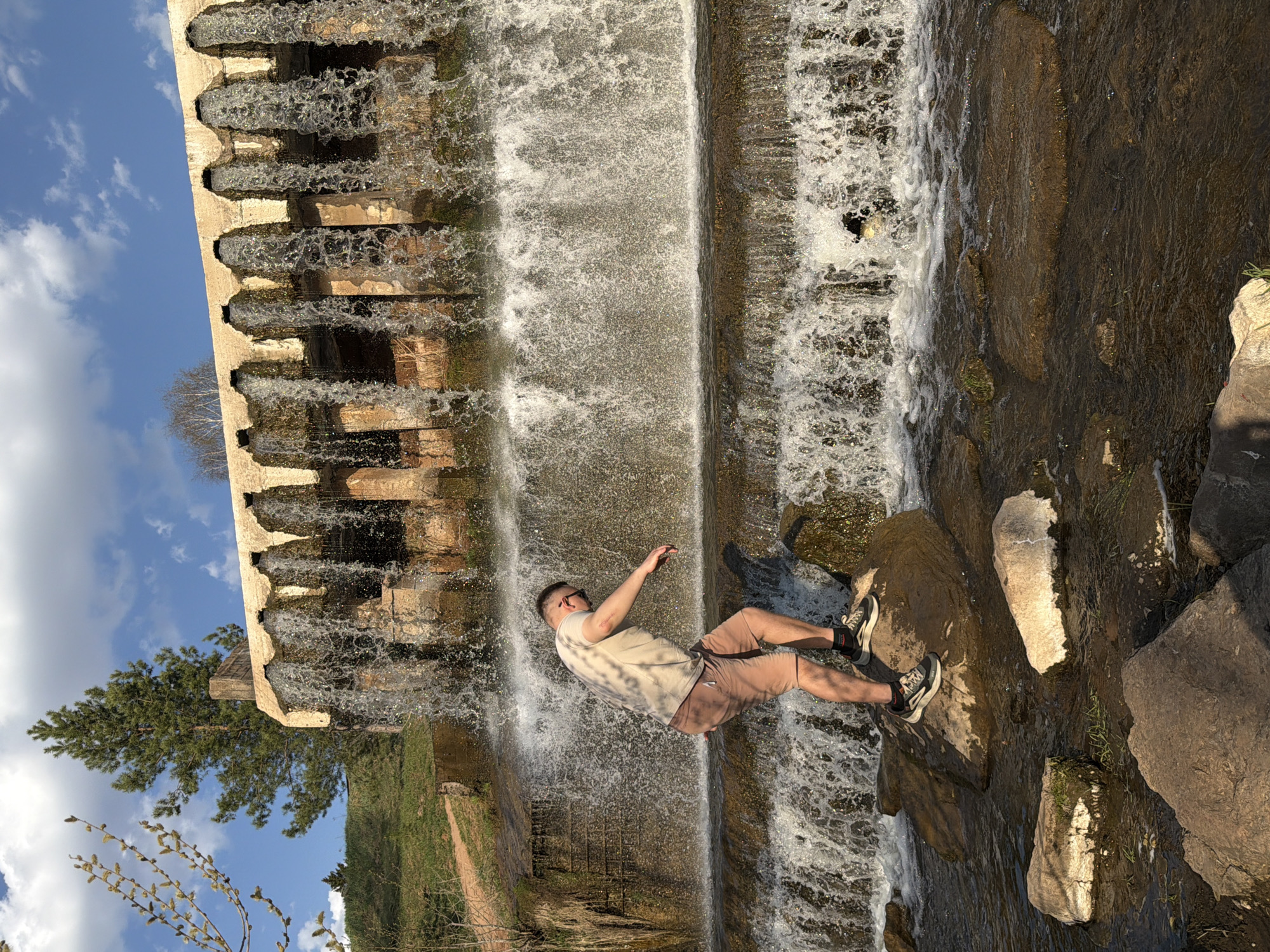 A man stands on rocks in front of a waterfall, which is part of a structure with multiple arched outlets. The location is Kamennoye, Russia. The man is wearing a light-colored t-shirt, brown shorts, and sneakers. He has one arm raised. The waterfall is cascading down a series of levels. There are trees and greenery in the background, as well as a blue sky with clouds.