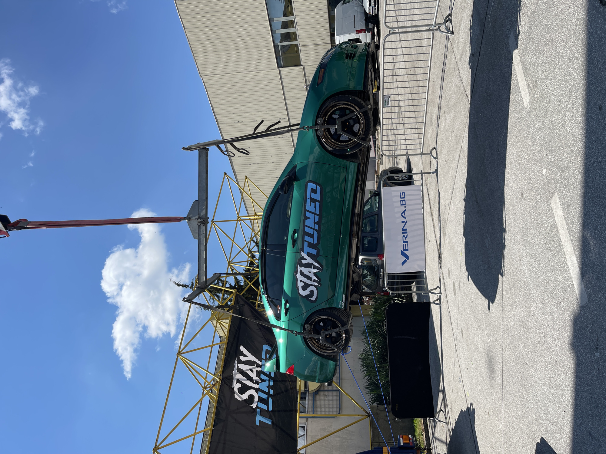 The image shows a green "STAY TUNED" customized car suspended in the air by a metal framework. The car is positioned between two signs: one black banner with "STAY TUNED" in white letters, and the other a white sign with "VERINA.BG" in blue. The scene is outdoors with a blue sky and a few white clouds visible. The setting appears to be a paved area, potentially at an event or exhibition in the city of Unknown, Unknown.