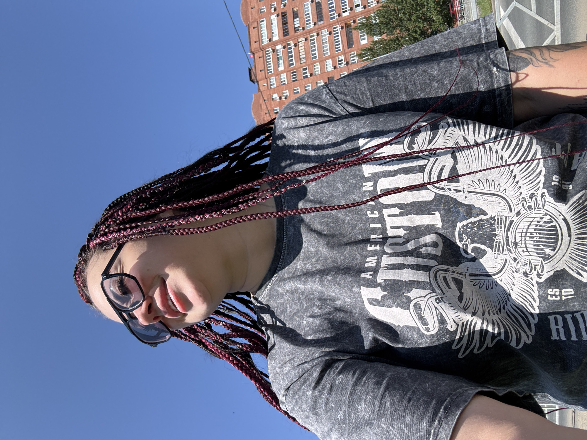 A young woman with auburn braids and black-rimmed glasses smiles as she looks to her left. She is wearing a gray t-shirt with a graphic print of an eagle and the words "AMERICAN CUSTOM". The background shows a bright blue sky and a multi-story brick building. The scene appears to be outdoors, possibly in the city of Tolyatti, Russia, given the architecture.