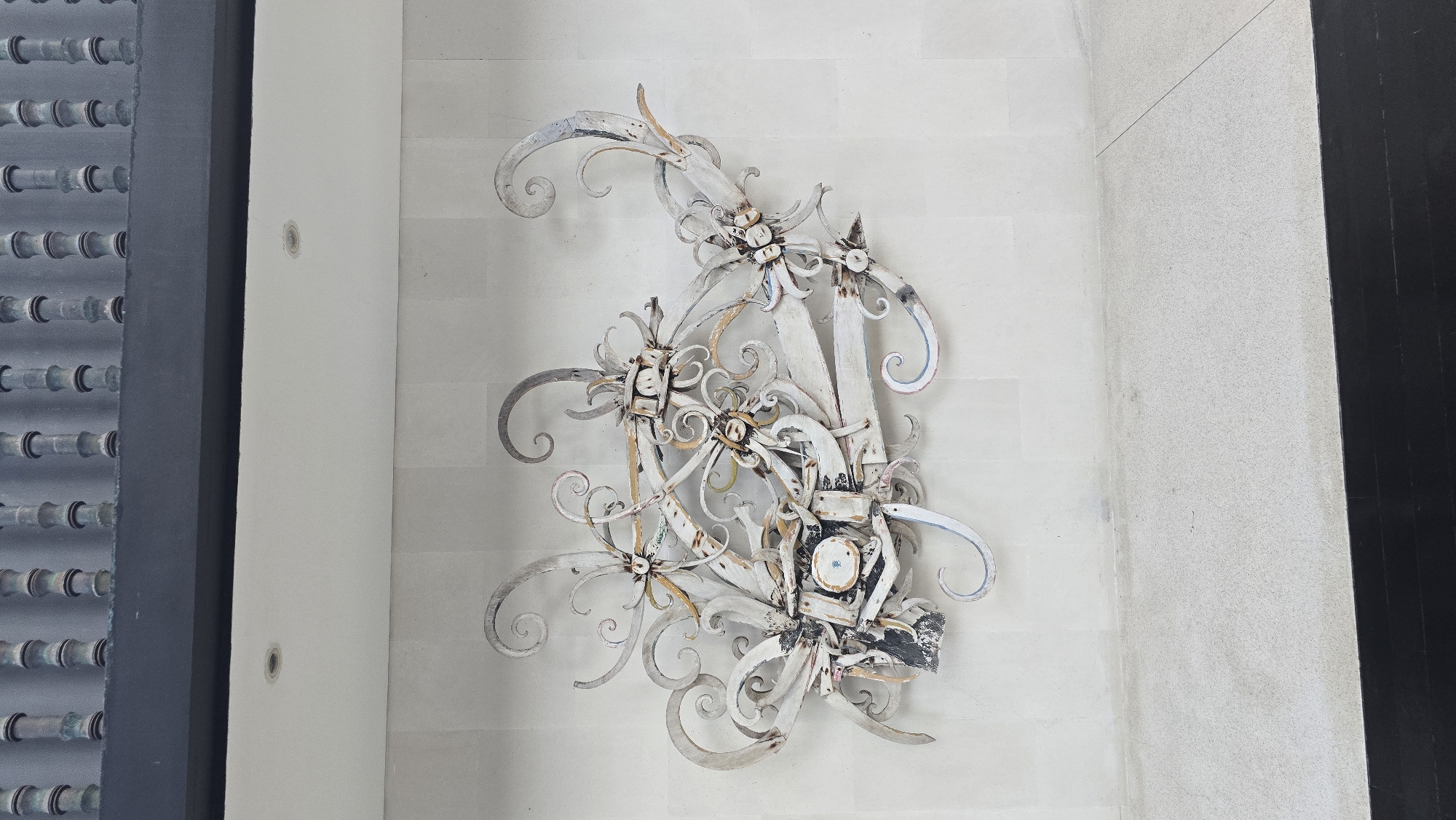 A close-up, slightly high-angle shot displays an ornate, wrought-iron decorative piece mounted on a textured white wall. The artwork has a swirling, floral-like design with multiple curls and stylized flower motifs. It is primarily white with hints of gold, gray, and blue. The texture suggests weathering and age. To the left, a vertical, gray panel with evenly spaced circular elements is visible. To the right, a dark, vertical surface defines the edge of the frame. The environment appears to be an interior space in Kuta Selatan, Indonesia.