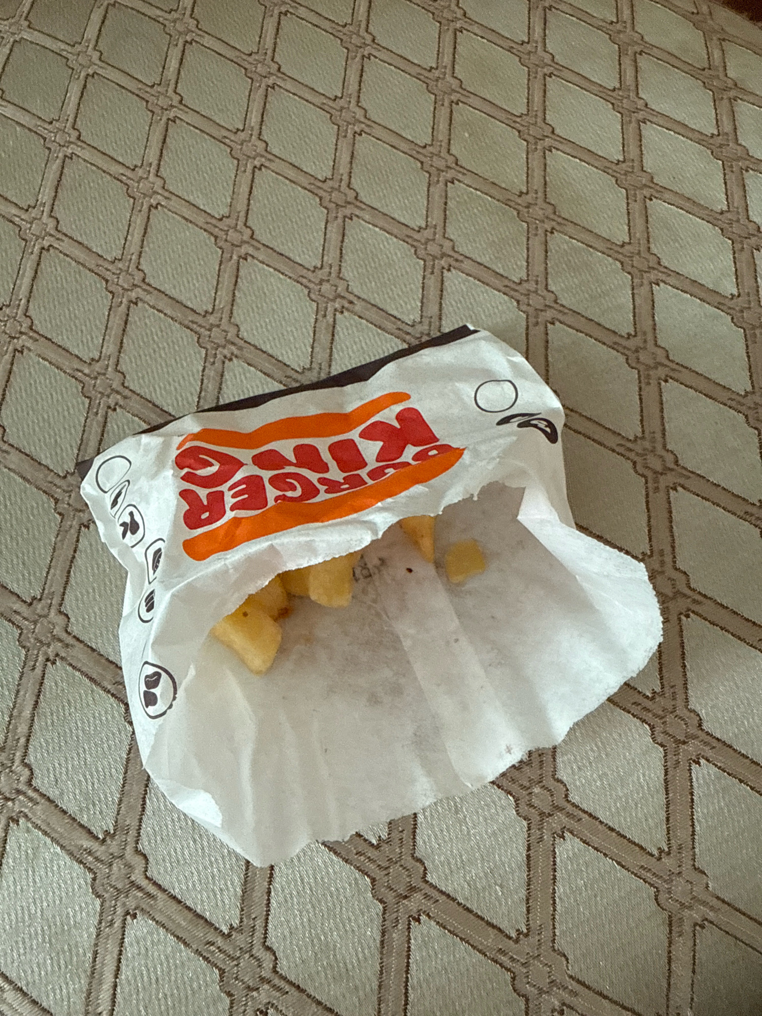 A partially opened Burger King paper bag containing french fries lies on a textured beige surface with a diamond pattern. The Burger King logo is visible on the bag. The image is taken from a top-down perspective.
