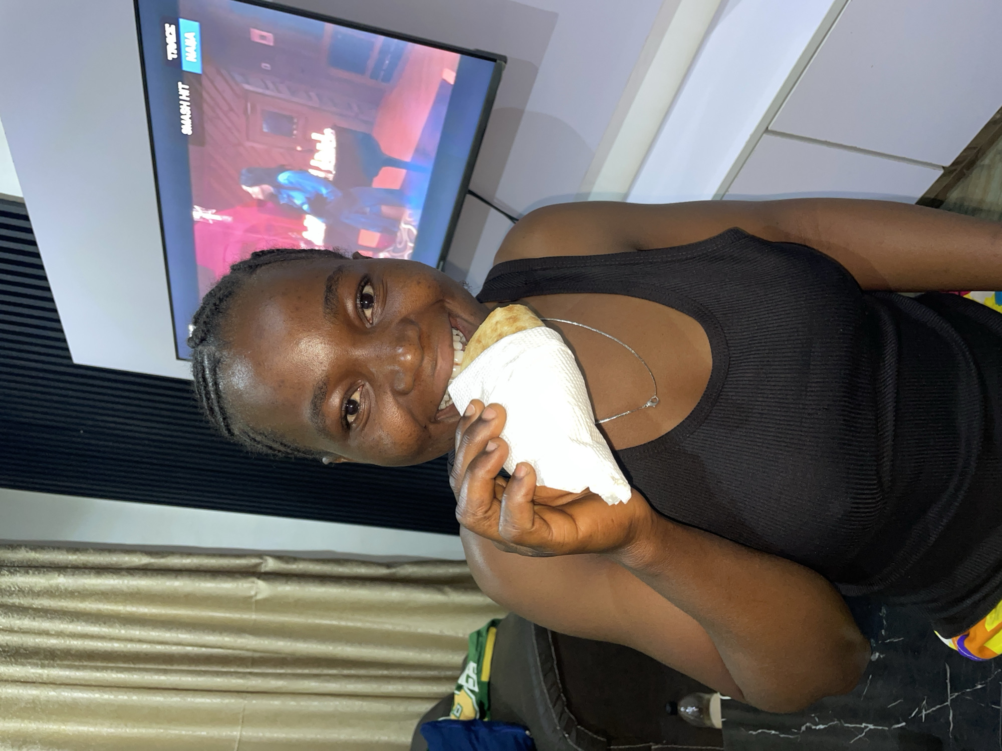 A dark-skinned woman with braided hair, wearing a black tank top and a silver necklace, is holding a food item wrapped in a white napkin. She appears to be taking a bite of the food. A television screen is visible in the background, displaying video game footage. The scene appears to be indoors, inside a home with curtains in the background and it is likely the city of Jos, Nigeria given the location of Jos, Nigeria.