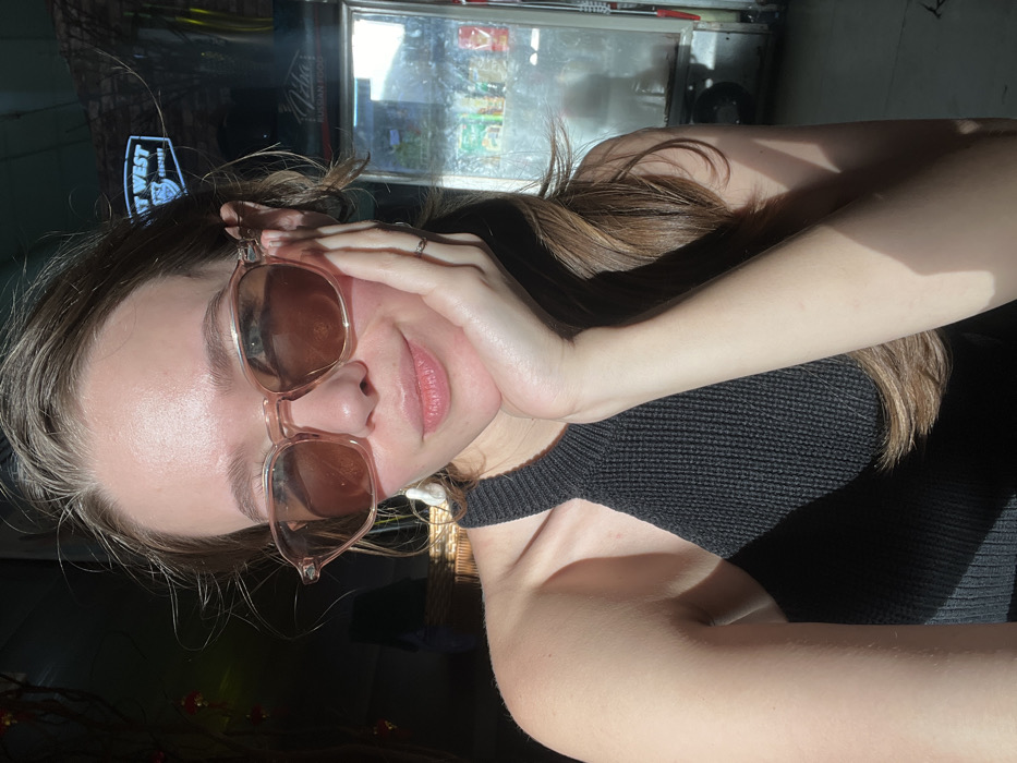 A fair-skinned female, appearing to be a young adult, is depicted from the chest up, positioned centrally in the frame. She wears light pink-framed sunglasses with brown tinted lenses and a black knitted halter-neck top. Her light brown hair is visible, extending over her right shoulder. Her left hand is raised, with fingers resting beneath her chin and along her jawline. A silver-colored ring is present on her left ring finger. She displays a slight smile and looks directly at the camera. Strong direct sunlight illuminates the right side of her face, shoulder, and arm, casting prominent shadows.
In the background, to the left, partially visible red decorative lanterns hang, alongside a dark, woven object. To the right, a commercial glass-front refrigerator is visible, containing various packaged items. Above the refrigerator, a sign is partially visible displaying "WEST WEST" and the explicit text "Petra RUSSIAN FOOD." The setting is an indoor or semi-covered area within a commercial establishment, likely a restaurant or cafe. This combination of details, particularly the "RUSSIAN FOOD" signage, indicates the location is in Nha Trang, Vietnam, a city known for its Russian tourist and expatriate community.